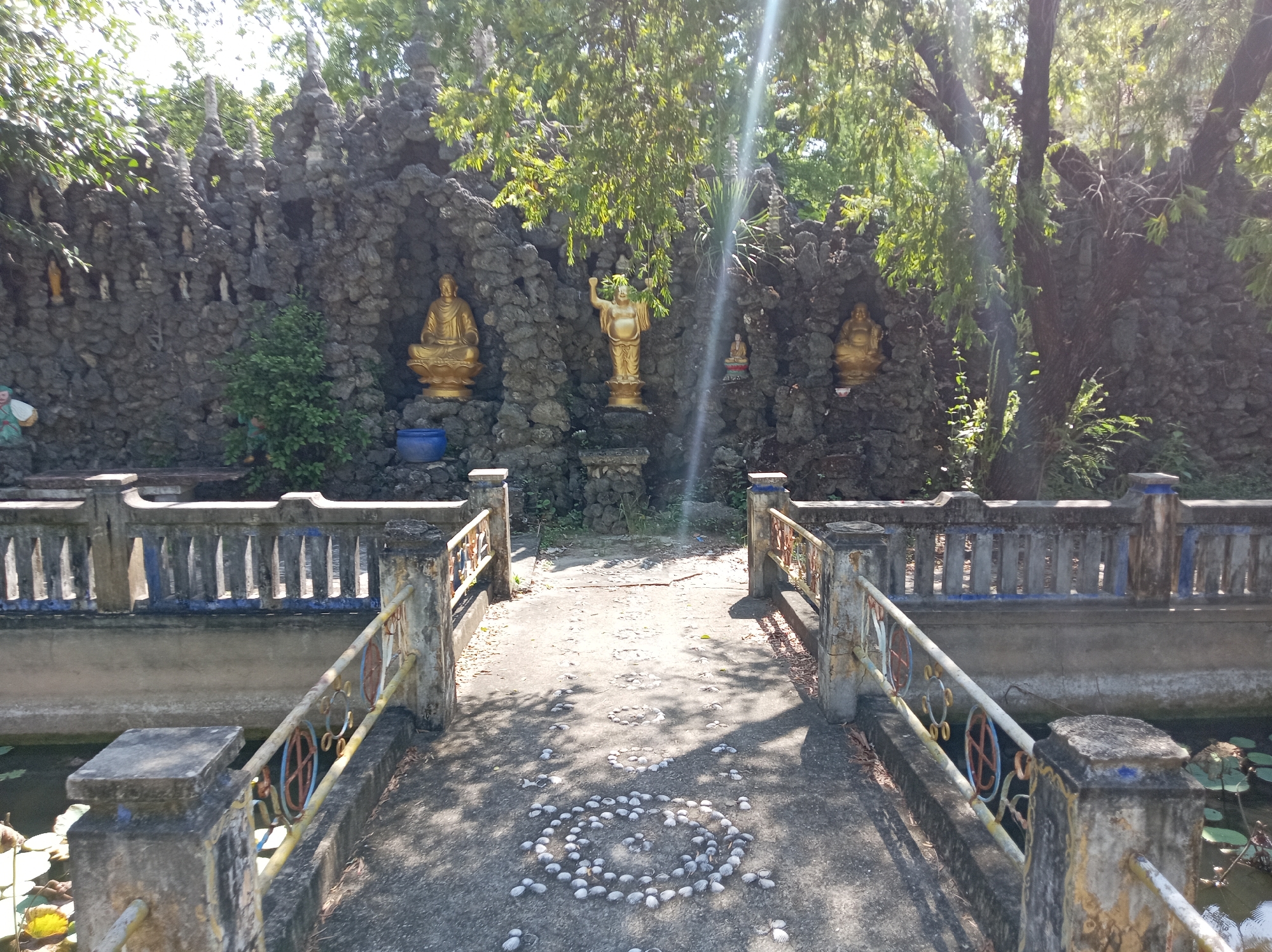 The image depicts an outdoor scene at a garden or temple located in Cam Ranh, Vietnam. A concrete pathway leads across a bridge-like structure, flanked by railings. The pathway is decorated with circular arrangements of shells. In the background, a wall constructed of dark, rough stones rises, with several gold-colored Buddha statues placed in niches. Trees with green foliage hang over the wall, and sunlight filters through the leaves, casting shadows on the ground. The environment appears to be tranquil and serene.