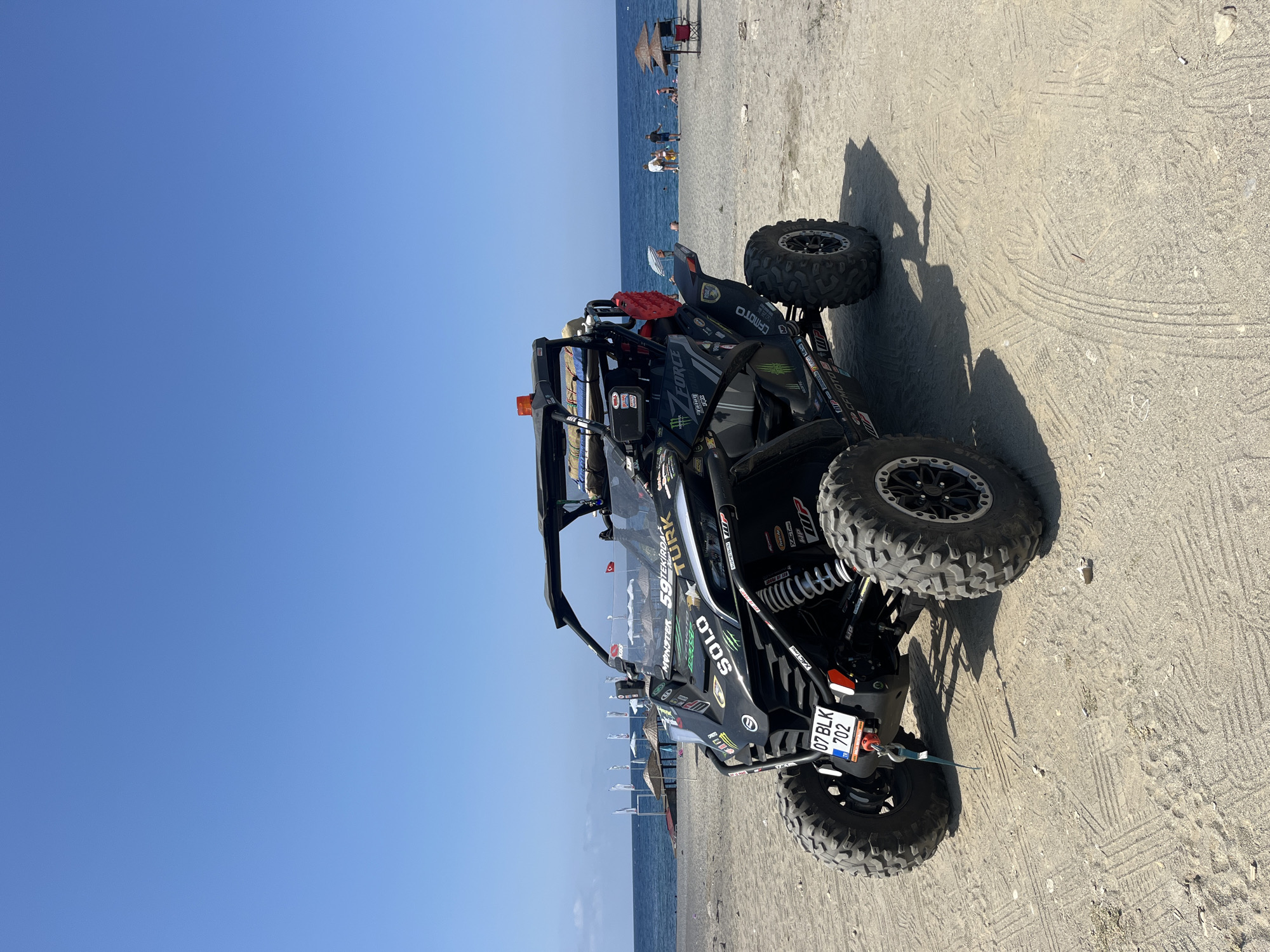 A black off-road vehicle, commonly known as a UTV or side-by-side, is parked on a sandy beach in Alanya, Turkey. The vehicle is heavily customized with decals and accessories, including sponsorships from brands like "MONSTER" and "7 FORCE". The front license plate reads "07 BLK 702" and displays the Turkish flag. The vehicle is positioned facing slightly to the left, with its large, knobby tires digging into the sand.
The background reveals a clear blue sky and the calm blue waters of the Mediterranean Sea. A few beach umbrellas and lounge chairs are visible along the shoreline, with a small number of people scattered across the sand, some wading in the water and others relaxing on the beach. The beach is composed of light-colored sand with visible tire tracks from the UTV. The overall scene suggests a recreational setting on a sunny day in Alanya.