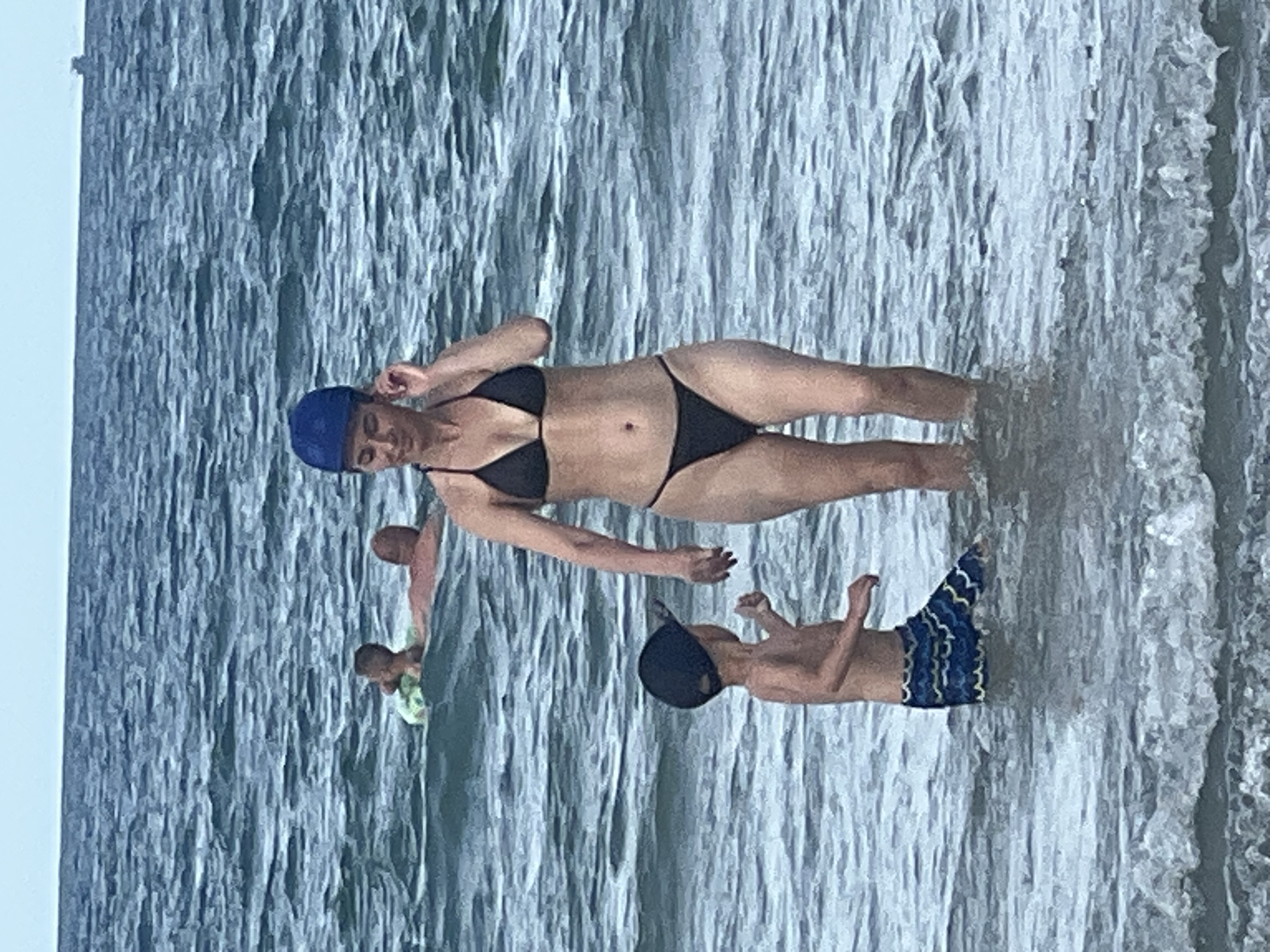 The image shows a scene at a beach in Da Nang, Vietnam. In the foreground, a woman in a black bikini and a blue cap is standing in shallow water. She appears to be posing or interacting with the water. To her right, a young boy wearing a dark blue cap and blue swim trunks with a patterned design is also in the water. He is facing the woman and seems to be engaged with her. Further in the background, another person is visible, partially submerged in the water, wearing a light green top. The water is wavy and appears to be a cool blue-grey color, suggesting a typical beach day. The lighting indicates it is daytime, likely a sunny day given the setting. The overall atmosphere appears relaxed and recreational, with people enjoying time at the beach. There is no visible text in the image.