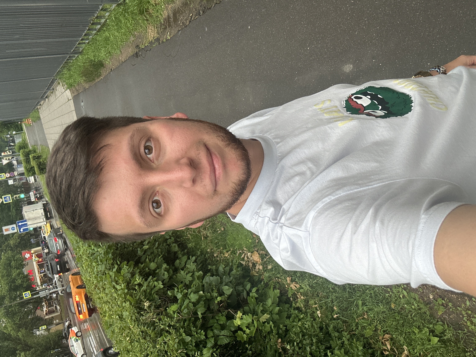 The image depicts a young man taking a selfie outdoors in Moscow, Russia. He is standing on a paved sidewalk next to a lush green hedge and a grey metal fence. The man is wearing a white t-shirt with a cartoon character graphic. Behind him, a street scene is visible with various vehicles, including a yellow convertible, a silver sedan, and a white truck, suggesting the image was taken in a developed urban area. The road appears wet, indicating recent rain. Traffic lights and street signs are visible, further confirming the urban setting.