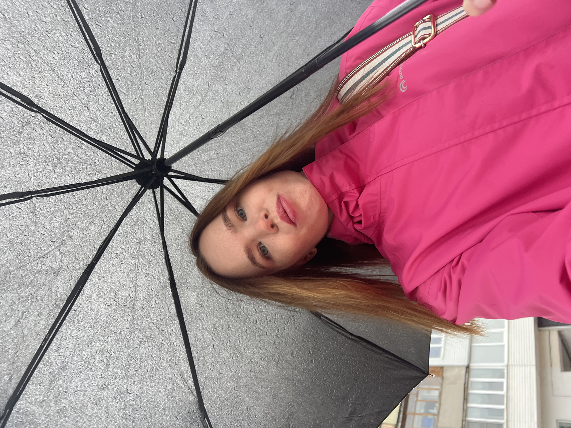 A woman is holding an umbrella outside in Chaikovsky, Russia. The umbrella is black with a gray, rain-spotted canopy. She is wearing a bright pink jacket with a striped strap crossing her chest. Buildings can be seen in the background.