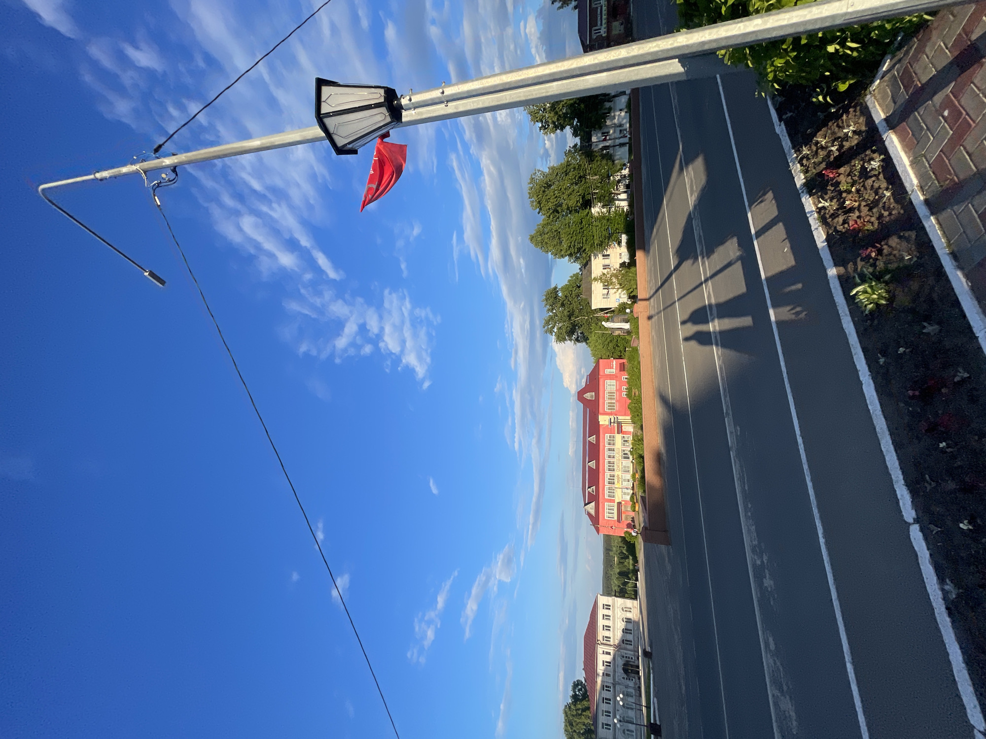 The image depicts a street scene in Krasnye Baki, Russia, under a bright blue sky with scattered white clouds. In the foreground, a paved road with white lane markings is visible, leading towards a row of buildings. To the right, a tall lamppost with an ornate black lantern stands prominently. A small red flag is attached to the lamppost, fluttering in the breeze.
In the middle ground, several buildings line the street. On the left, a long, light-colored building with multiple windows and an arched entrance is present. To its right, a larger, brightly colored pink building with a red roof and numerous windows dominates the scene. Further back, more buildings and green trees can be seen, suggesting a developed urban or semi-urban area.
The shadows of two individuals are cast on the road, indicating the presence of people, though they are not directly visible. The overall impression is a sunlit day in a town square or along a main street.