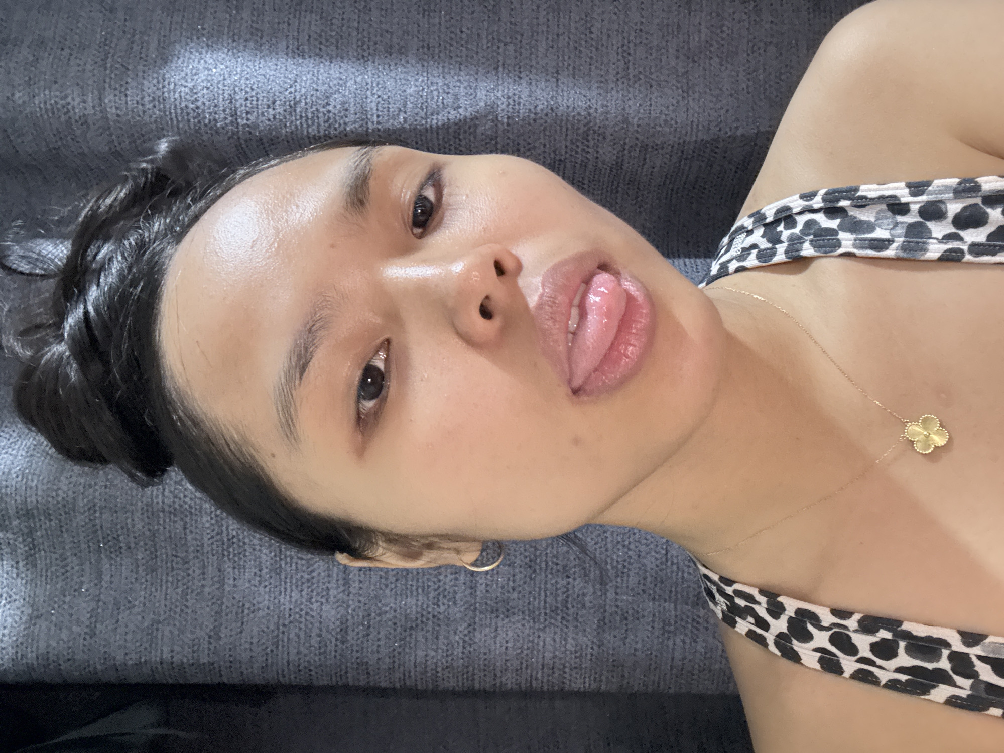 A woman lies on a gray, textured surface. She has dark hair pulled back in a bun and is wearing a top with a black and white spotted pattern. The woman's tongue is slightly extended. She is wearing a gold necklace with a clover-shaped pendant. A small gold hoop earring is visible. The location is not specified in the image or hints; therefore, there is no information to suggest that the image was taken in Davao City, Philippines.