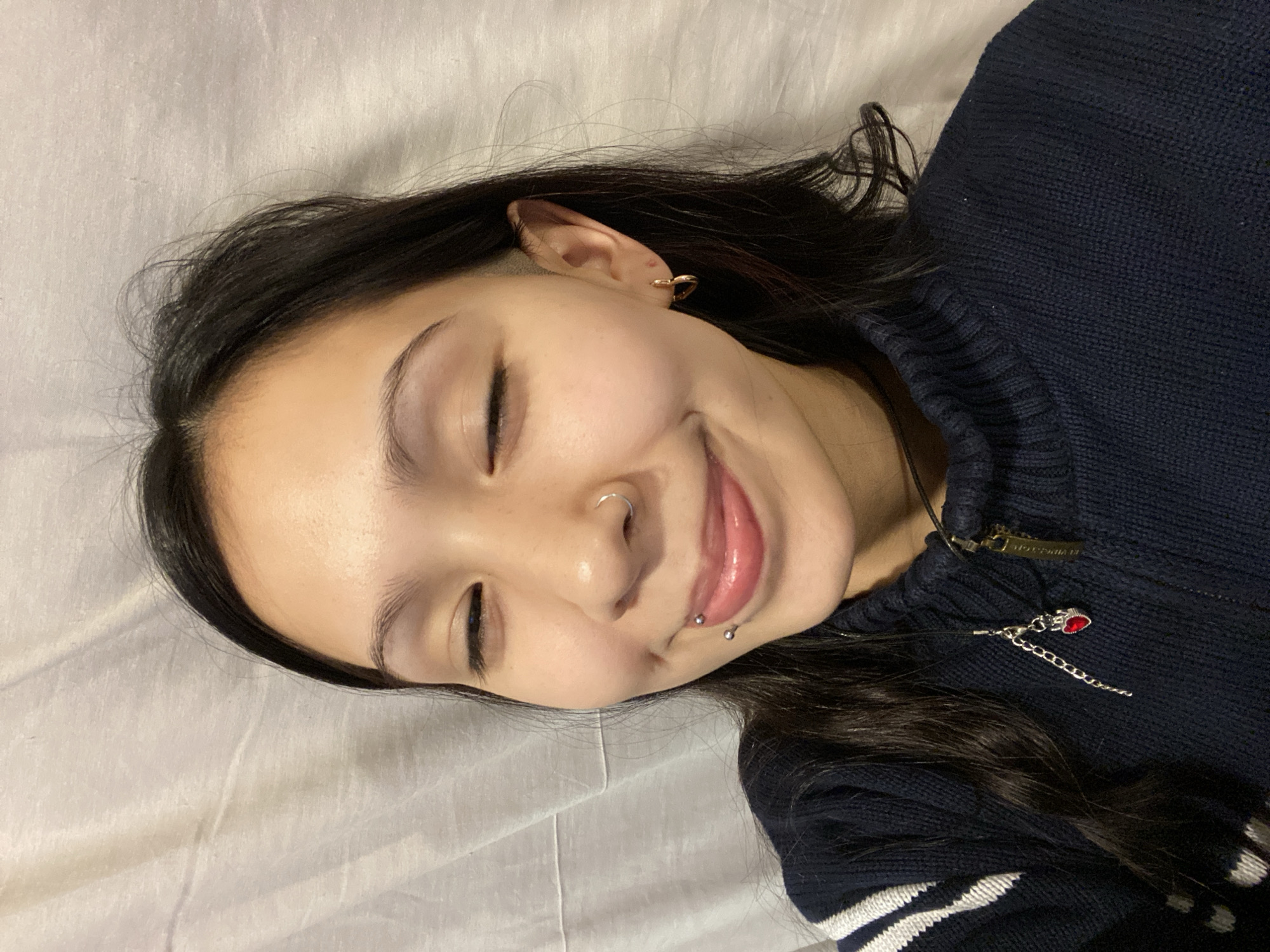 A young woman lies on a white fabric surface, possibly a bed or soft floor, angled towards the viewer. Her dark hair is parted to the side, with a portion appearing to be closely shaved near her ear. She has her eyes closed and a slight smile, revealing a lip piercing on the left side of her mouth. A small silver nose ring adorns her left nostril. She wears a navy blue knit sweater with white stripes visible on the lower right side, and a delicate silver chain necklace with a small red pendant is visible near her collarbone. The setting is indistinct, offering no clues as to its location within Buenos Aires, Argentina.