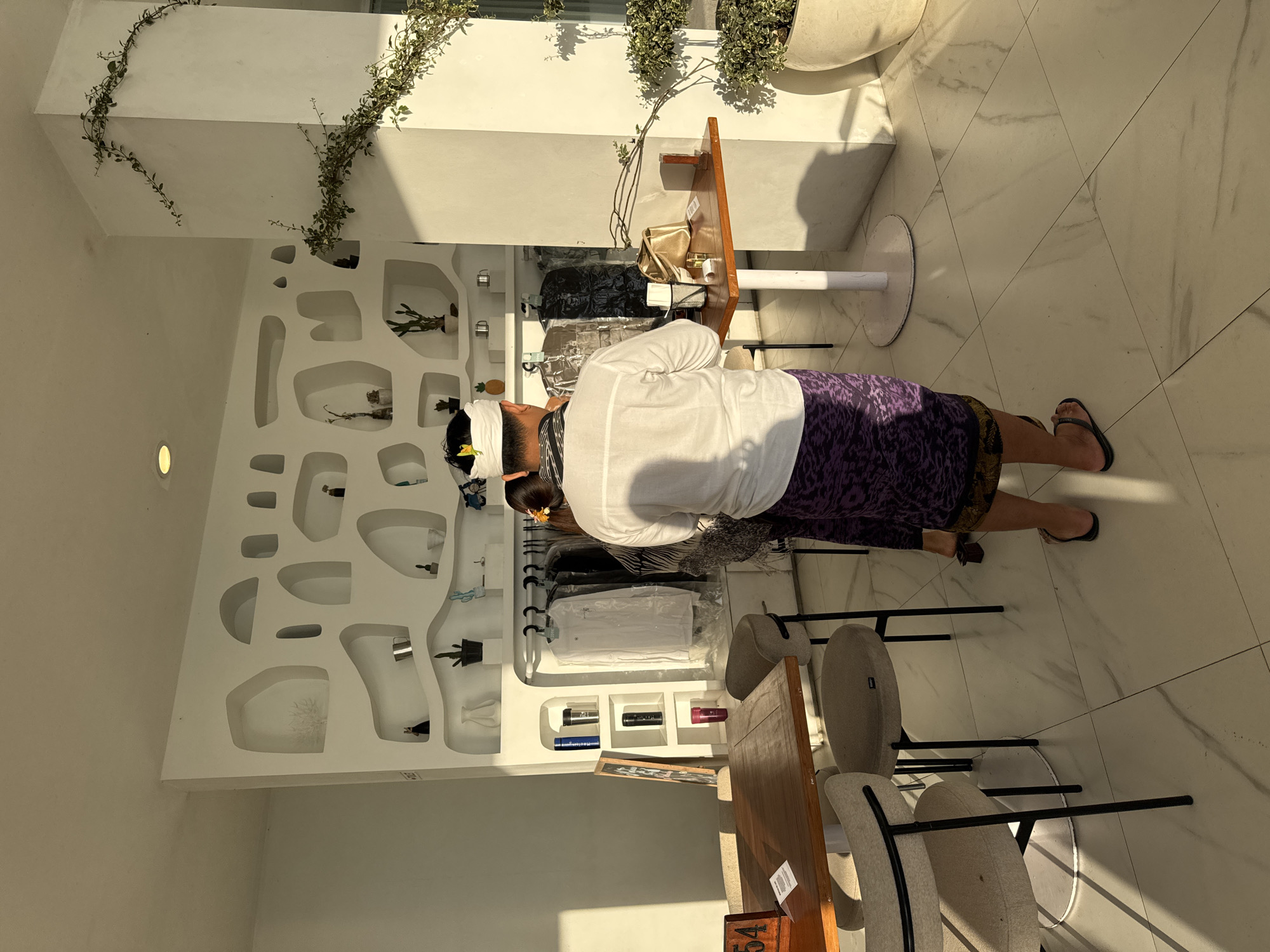 The image, taken in Kedisan, Indonesia, shows a man inside a retail space. He is wearing a white shirt, a purple sarong, and a traditional white headdress with a flower. He is wearing flip-flops. He appears to be examining items on a clothing rack inside the shop. The shop's interior is characterized by a white wall with built-in display niches, some containing decorative items. A wooden table with chairs stands in the foreground. Above, there is foliage decorating the space. The floor is light-colored tile.
