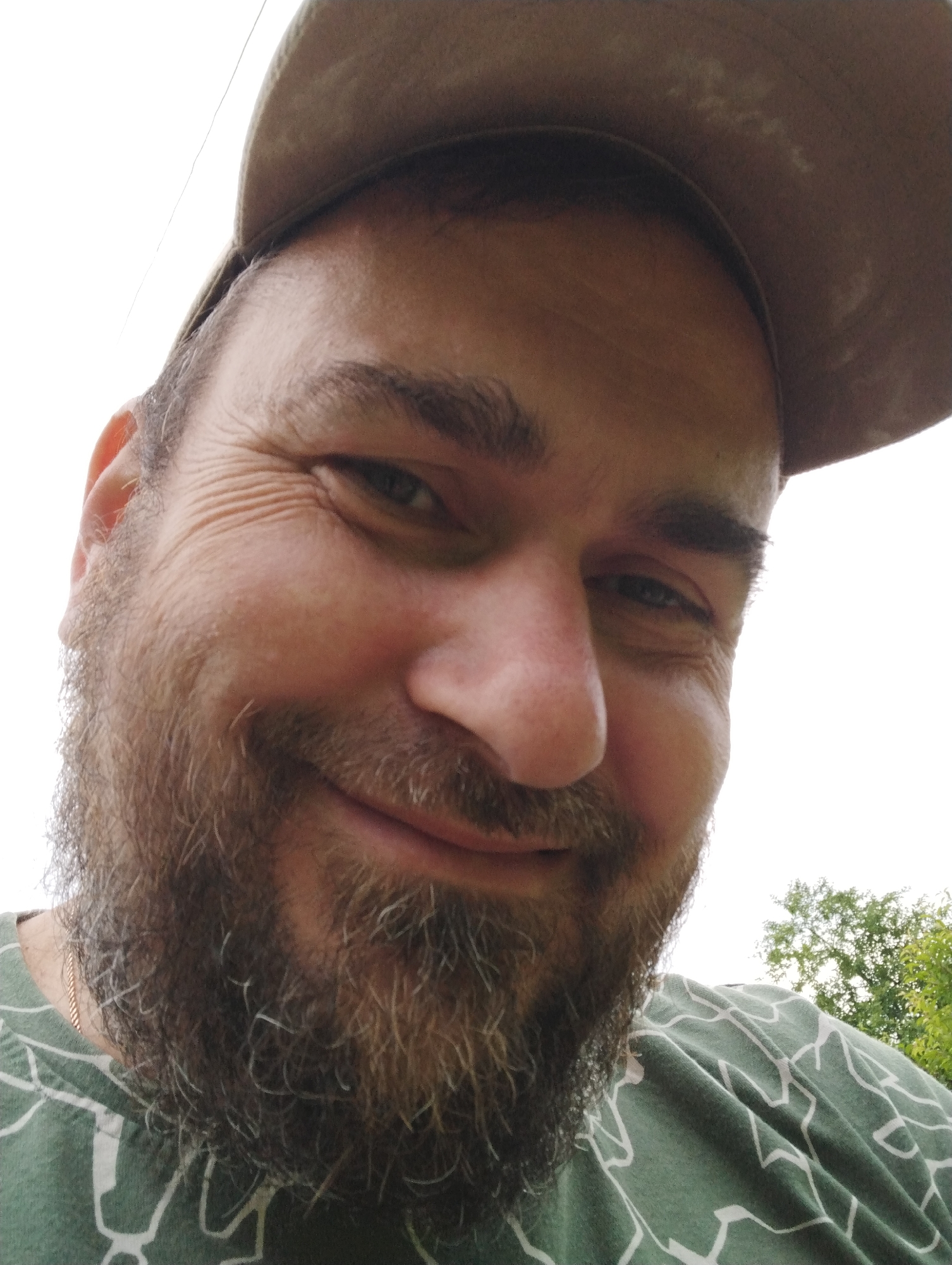 A close-up selfie of a man wearing a brown baseball cap and a green t-shirt with a white geometric pattern. He has a beard and is looking towards the camera with a slight smile. The background is out of focus, showing a bright sky and some green trees, suggesting an outdoor setting. The lighting indicates it is daytime. The exact location in the city of Ширяево (ильинский), Russia cannot be determined from the image alone.