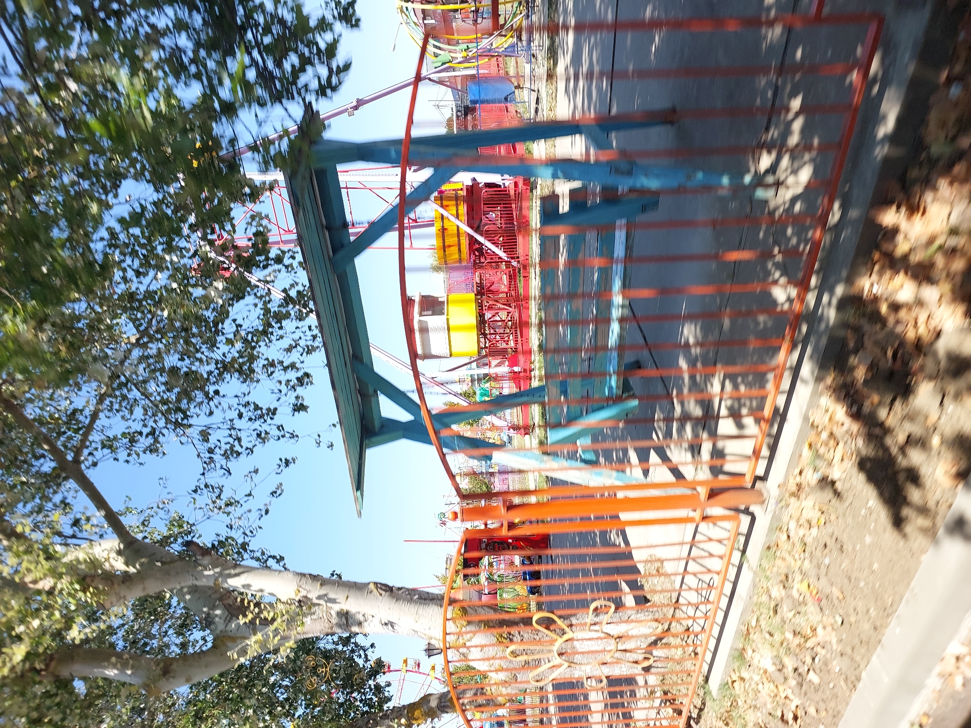 An outdoor amusement park scene in Vityazevo, Russia, is depicted. A large tree with light-colored bark and green leaves is prominently featured in the left half of the frame, casting shadows. In the foreground, a red metal fence with vertical bars runs across the image. Behind the fence, a blue and teal colored swing set with a flat roof is visible. To the right of the swing set, parts of amusement rides with red, yellow, and blue components can be seen. In the background, a Ferris wheel with yellow cars is visible against a clear blue sky. The ground is covered with dry leaves and dirt. The image appears to be taken on a sunny day.