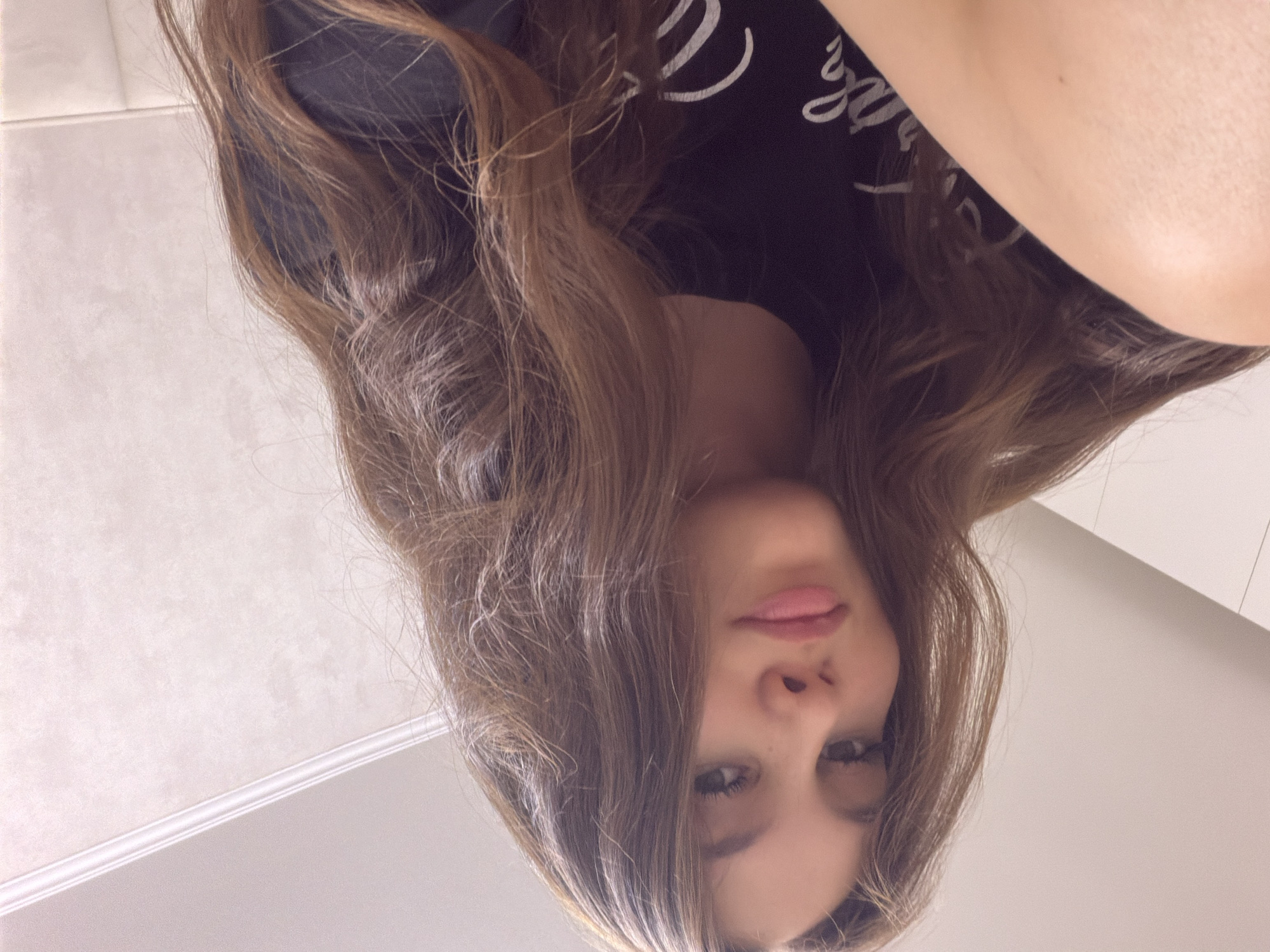 A close-up, low-angle selfie shows a young person with long, wavy brown hair and light skin. They are wearing a black t-shirt with white lettering. The person is looking towards the camera with a neutral expression. The background appears to be an indoor setting with a lightly textured wall and a white ceiling trim. The lighting is soft and diffused. There is no discernible indication of the location being in Naberezhnye Chelny, Russia, or any other specific city.