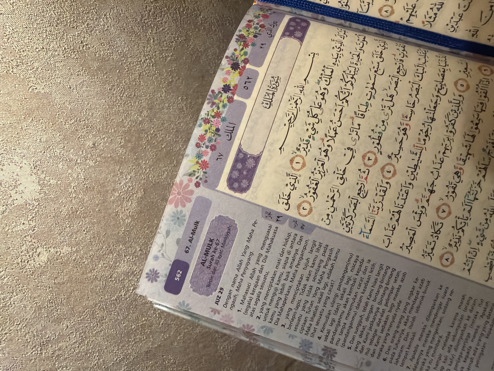 The image shows an open Quran, with the Surah Al-Mulk (Chapter 67) displayed. The text is in Arabic, with an Indonesian translation in the margins. The page is decorated with floral patterns and colored sections indicating the Juz (part) and Surah number. The image is shot close-up, focusing on the Quran itself.
