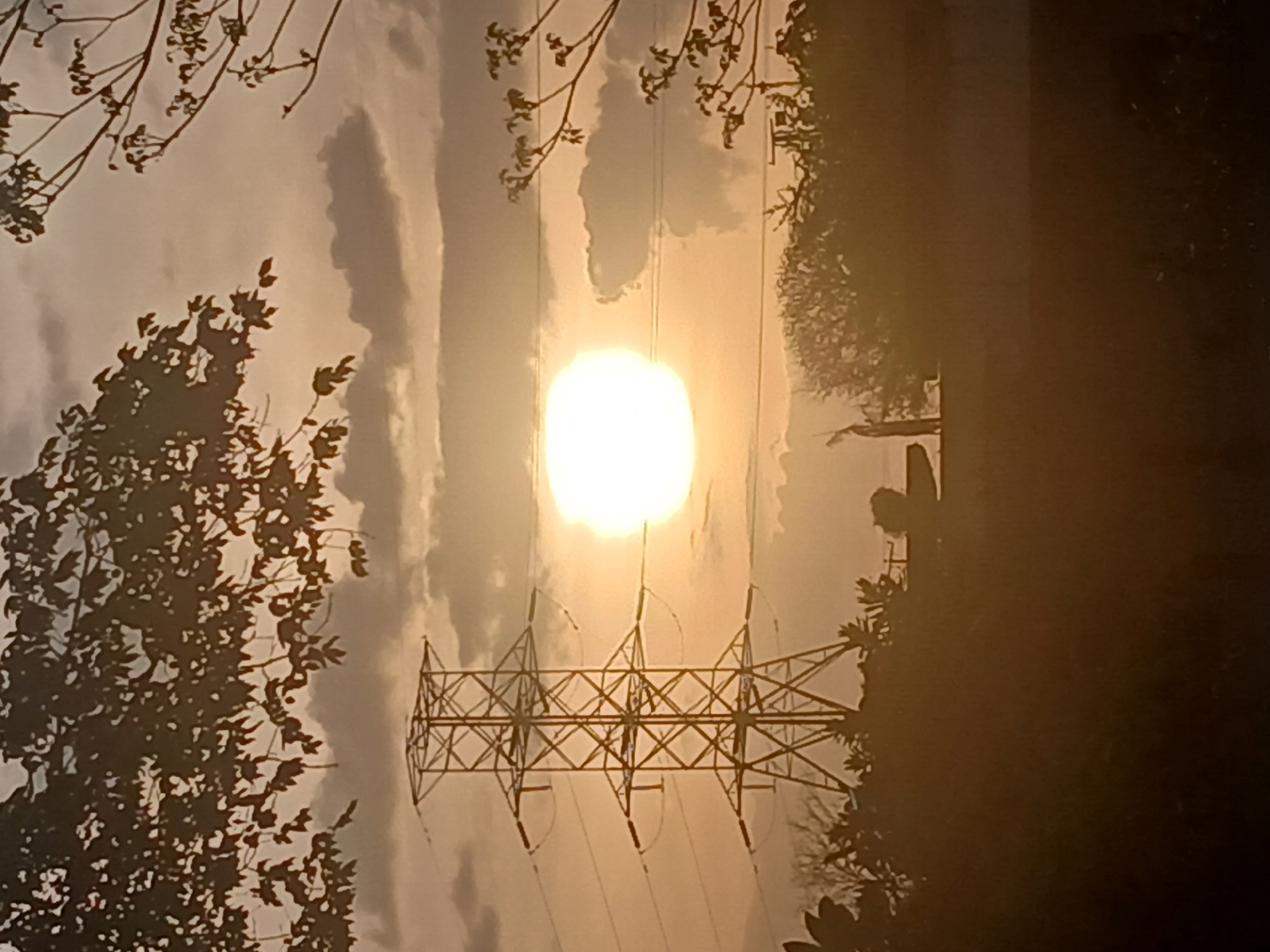 The image shows a power line tower silhouetted against a bright, pale sun or light source in a cloudy sky. Vegetation and trees are silhouetted on both sides of the tower. The scene appears to be outdoors, likely in an area where power lines are prevalent. There is no information in the image to confirm its location as Ciudad Guayana, Venezuela.