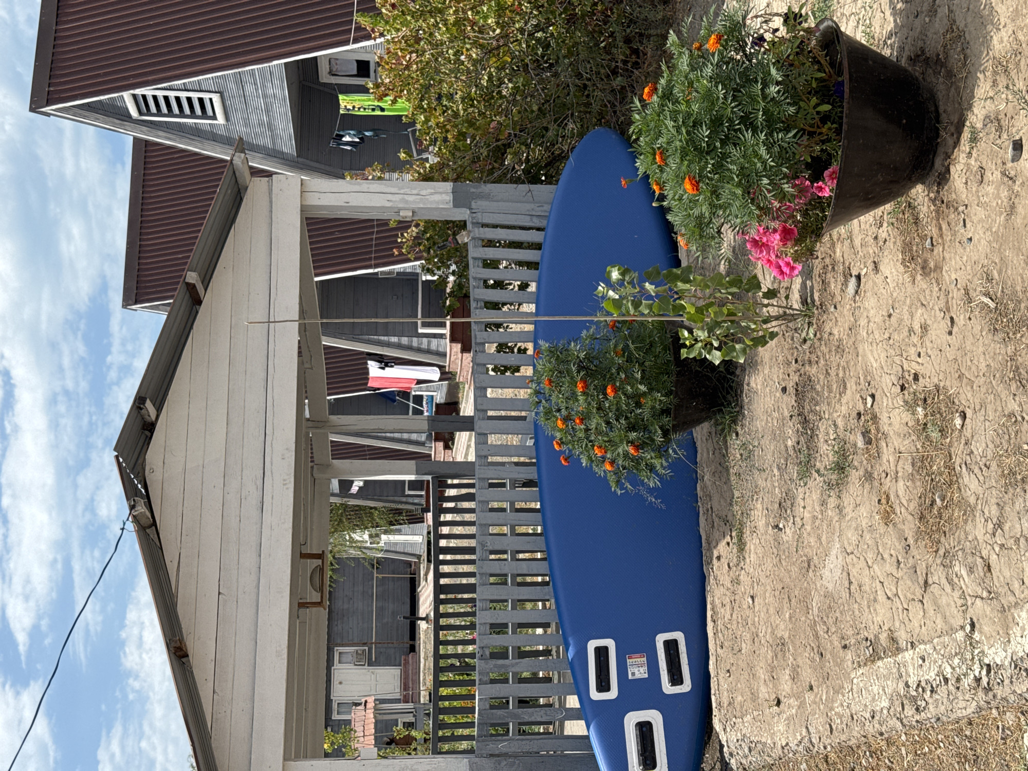 In Zharbulak, Kazakhstan, a blue surfboard lies on the ground in front of a gray wooden fence. Behind the fence, a white gazebo with a corrugated metal roof is visible. Further back, several triangular-roofed houses with brown metal roofs are arranged in a row, some with laundry hanging outside. Potted plants with orange and pink flowers are placed near the surfboard. The ground is covered in dirt and dry grass. The sky is blue with scattered clouds.