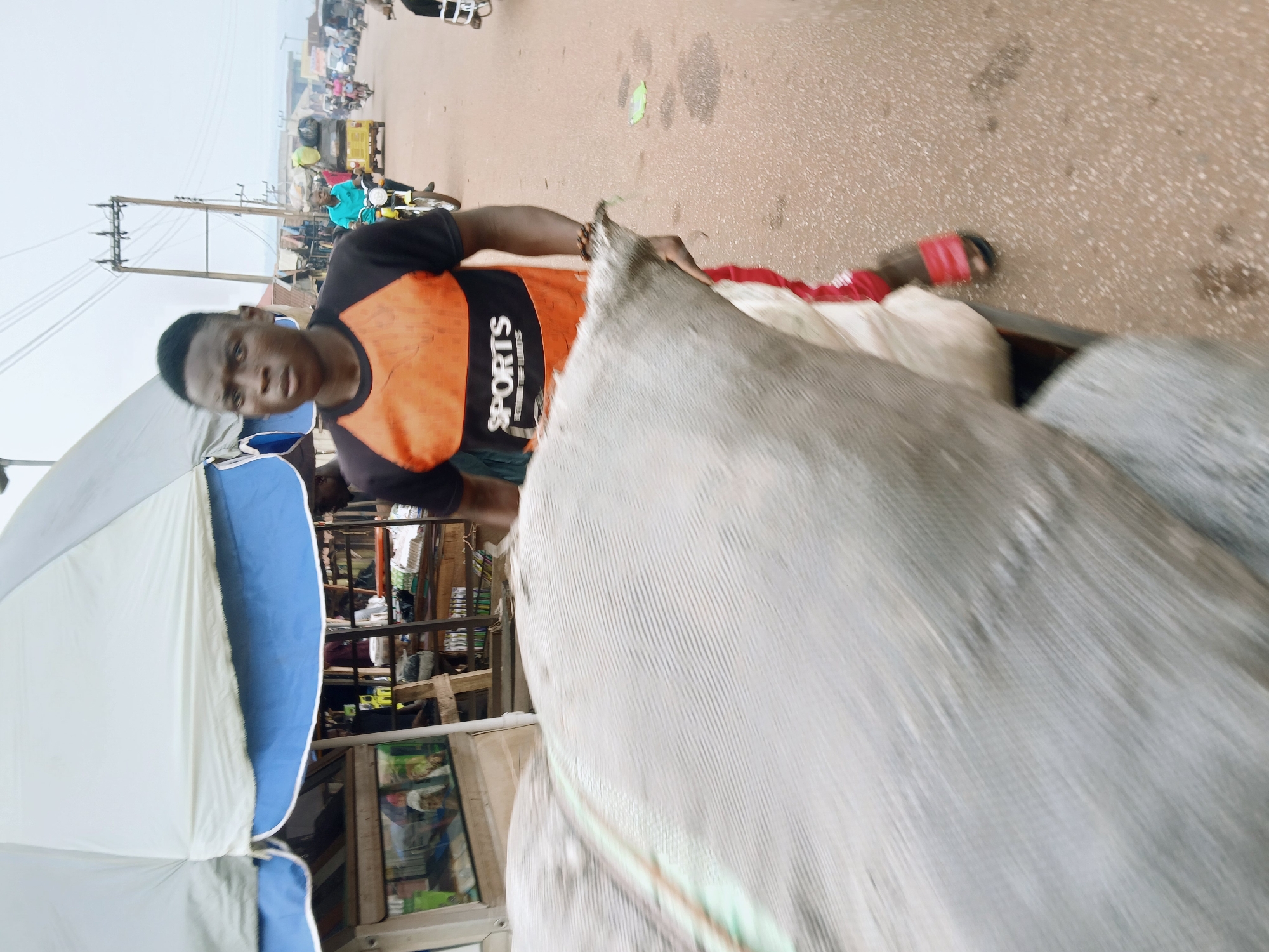 The image depicts a man, likely a vendor or seller, standing on a road in Ankpa, Nigeria. He is positioned behind a cart or vehicle laden with bulky sacks, possibly containing goods for sale. He is wearing a t-shirt with a black upper portion, an orange panel featuring the word "SPORTS" in white lettering, and dark shorts. The road behind him shows signs of vehicular and pedestrian traffic, including motorcycles and other vehicles. In the background are buildings and structures that appear to be part of a market or commercial area. An umbrella with white and blue panels provides some shade.