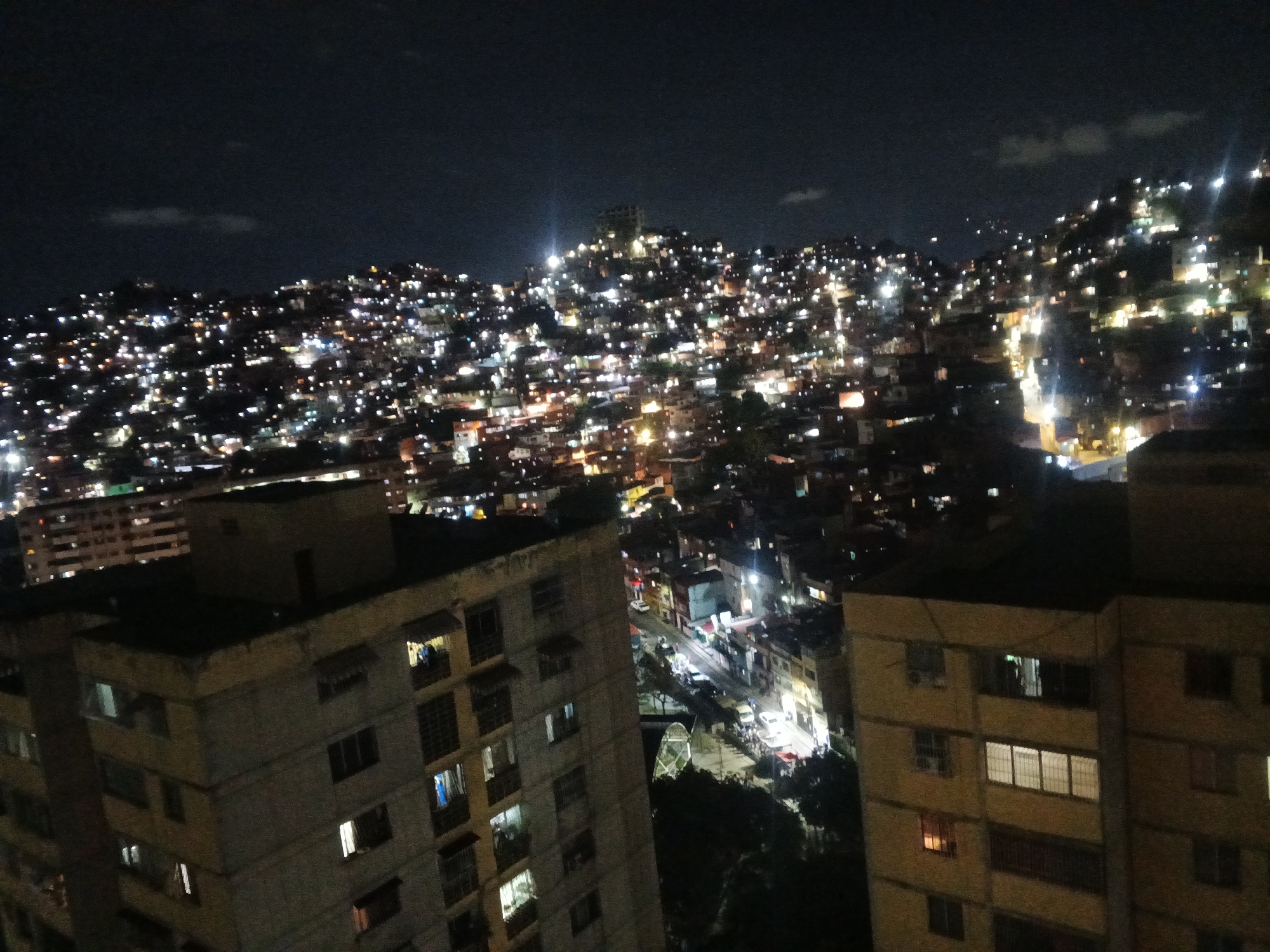 The image depicts a nighttime view of Caracas, Venezuela. The foreground shows two apartment buildings with visible windows. A brightly lit street runs between the buildings. In the background, a densely populated hillside is illuminated by numerous lights from homes and businesses. The sky is dark, with a few faint clouds visible.