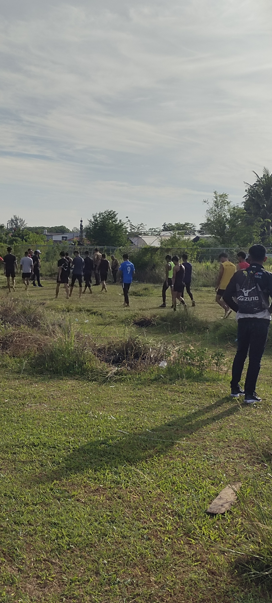 The image depicts an outdoor scene with a group of young men in a grassy field under a partly cloudy sky. The environment suggests a location in Banda Aceh, Indonesia, given the typical climate and presence of lush vegetation. In the background, there are trees, some buildings, and possibly a religious structure like a mosque.
The foreground is dominated by a well-maintained grassy area, with some patches of dry grass and a few scattered pieces of debris, including a small piece of wood. The lighting suggests it is daytime, likely late afternoon, as indicated by the long shadows cast by the individuals on the grass.
The main subjects are a group of approximately 15-20 young men, predominantly wearing athletic attire such as t-shirts, shorts, and athletic pants. Several are engaged in various activities, some appearing to be stretching, others conversing, and some are walking or standing around. One individual in the foreground, on the right, is wearing a black backpack with a prominent "MIZUNO" logo. Another individual in the middle of the frame is wearing a blue t-shirt with the number "9" on it. The group appears to be in a casual setting, possibly a sports practice or a gathering.