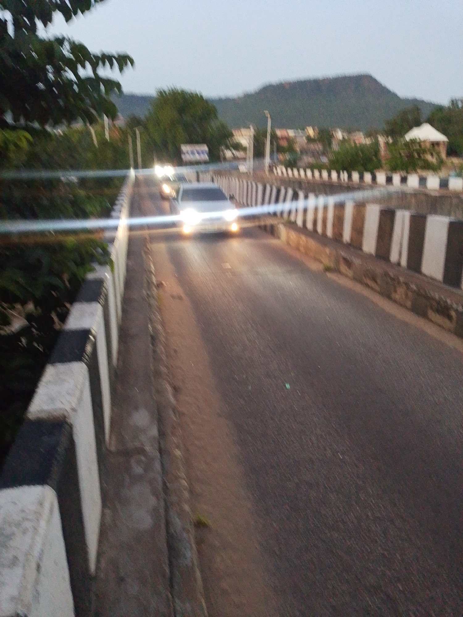 The image depicts a bridge with black and white striped barriers on either side. Cars are driving on the bridge with their headlights on. The surroundings include greenery, buildings, and a mountain in the background. The location is presumed to be a city, but the specific location is unknown.