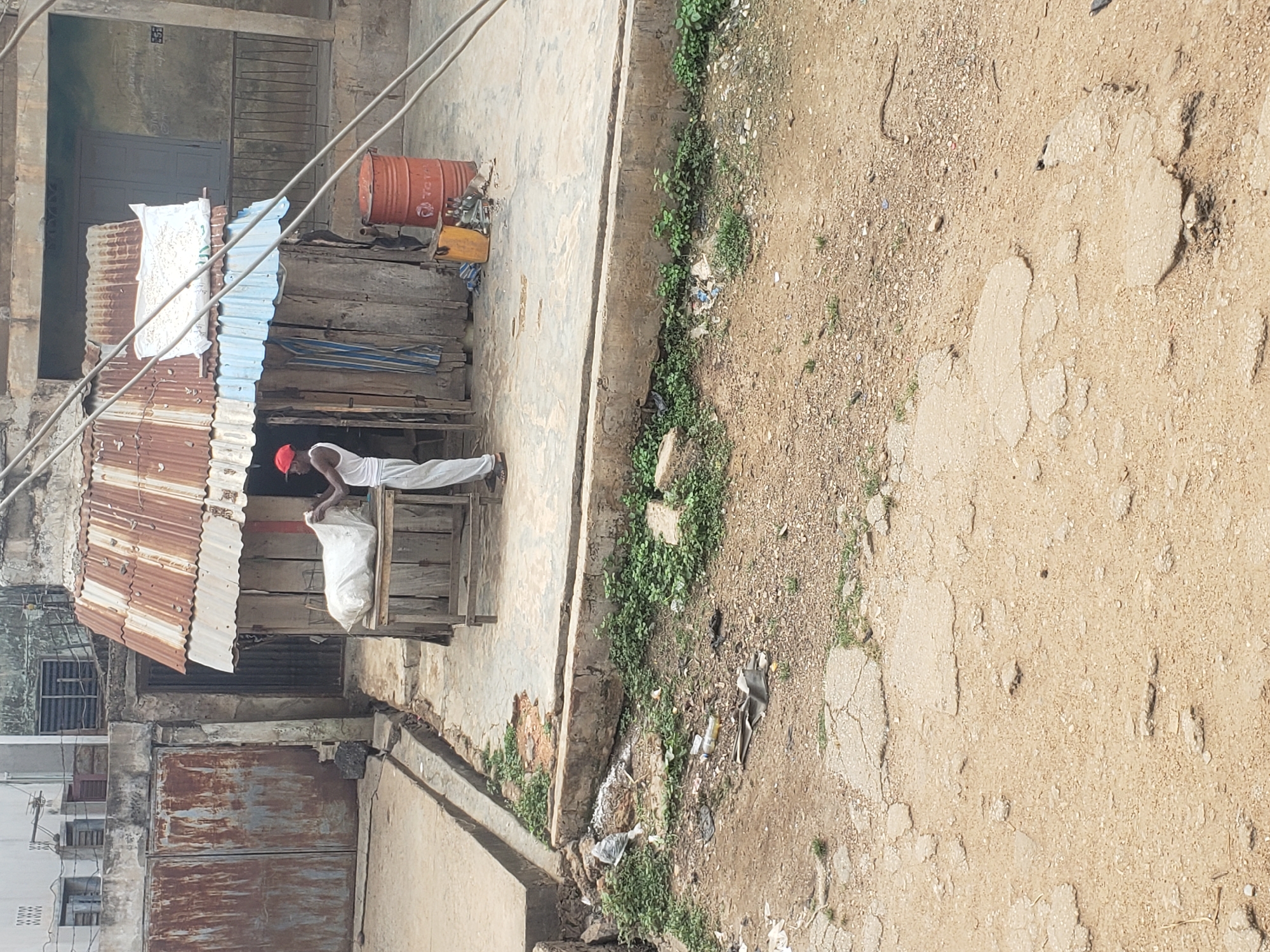 The image depicts an outdoor scene in what appears to be a local street or alleyway in Alalubosa, Nigeria, under bright daylight. The ground is a mix of dirt, gravel, and cracked concrete patches, with sparse green vegetation growing along edges and in crevices.
In the center-left of the frame, one individual is visible, dressed in a white sleeveless top and light-colored trousers, wearing a red cap. The person is positioned in front of a small, rustic structure made predominantly of corrugated metal sheets and wooden planks, which appears to be a makeshift stall or shop. The individual is engaged in handling a large, white sack, possibly arranging goods or making a sale.
Behind the person and the stall, there are older, concrete buildings with weathered walls and some visible windows, indicating a built-up area. Overhead, utility wires crisscross the scene. To the right of the stall, an orange metal barrel, possibly a fuel drum with "TOTAL" visible on its side, rests on a wooden platform. Nearby are other miscellaneous items and some debris on the ground. The overall impression is one of a bustling, informal commercial setting in a local neighborhood.