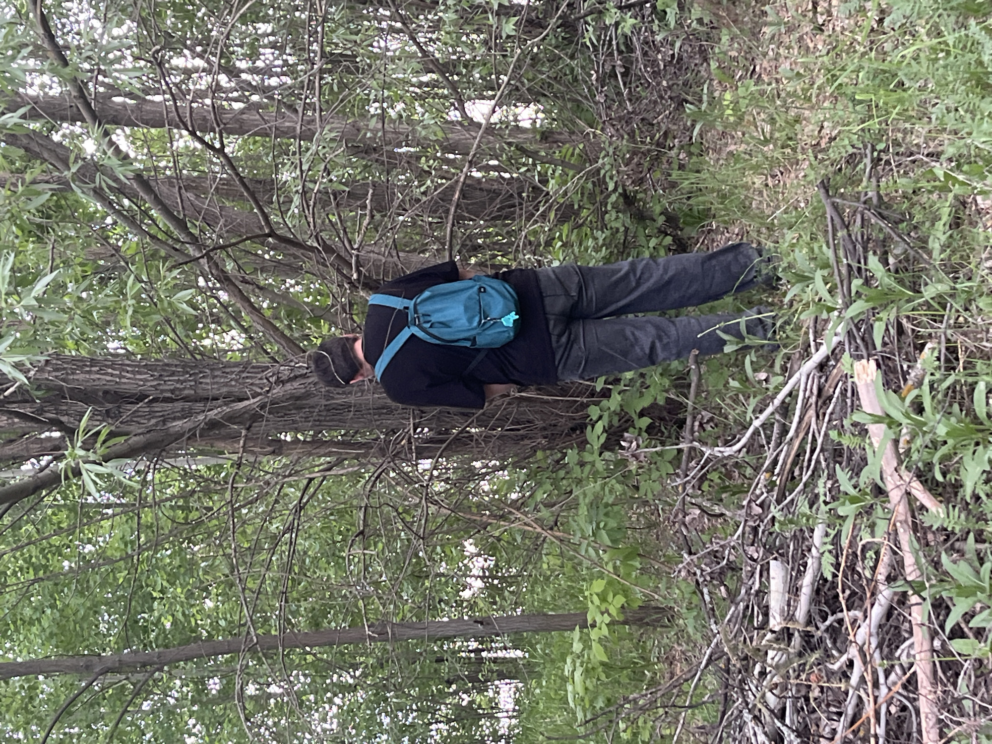 A man in a black shirt, dark pants, and a dark baseball cap is standing in a wooded area. He has a blue backpack on. The scene appears to be located within a densely wooded area, with trees and ground vegetation visible. The location is described as Chaikovsky, Russia, suggesting an outdoor environment with natural features consistent with the region.