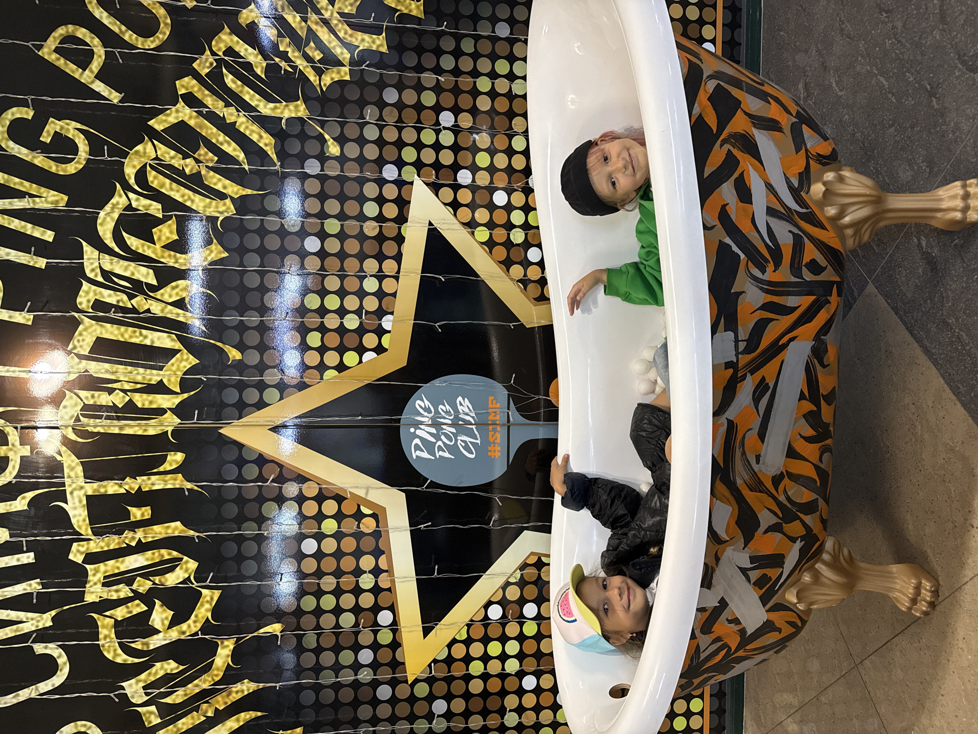 Two young girls, one wearing a watermelon-patterned hat and a black jacket, and the other wearing a black beanie and a green hoodie, are sitting in a white bathtub. The bathtub is decorated with an orange, black, and gray abstract graffiti pattern and rests on ornate gold legs. The background features a black wall with gold gothic lettering that reads "PING PONG CLUB." A large gold star is superimposed over a wall of shimmering circles. The scene appears to be an indoor, possibly recreational, setting in an unknown city.