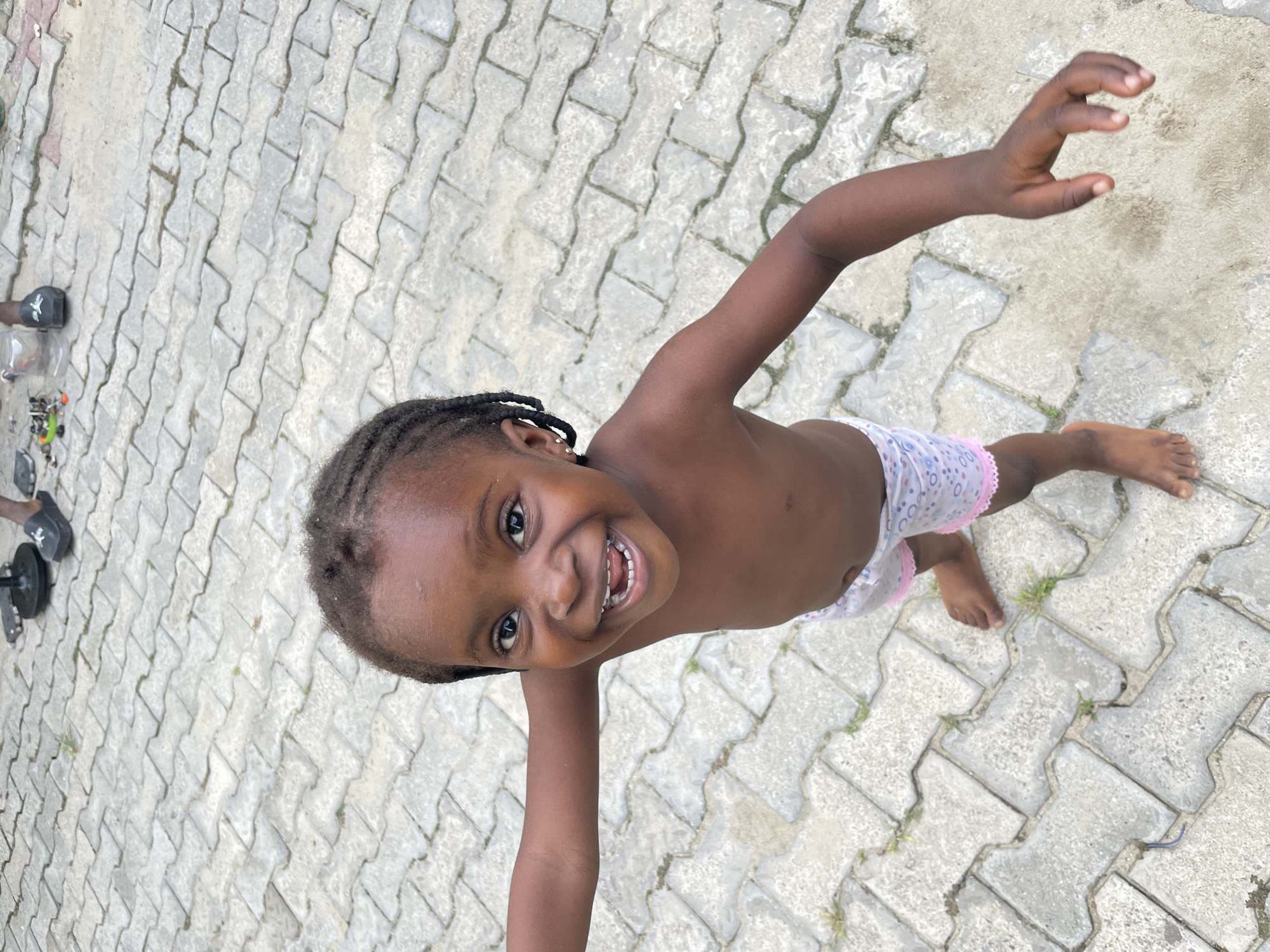 A young, dark-skinned girl with braided hair stands on a gray brick patio, facing the camera with a broad smile. She has a gold stud earring in her left ear. She wears patterned shorts, and her upper body is bare. Her arms are outstretched, and one foot is slightly raised. The patio surface appears to be made of interlocking pavers. In the background, a pair of sandals and a few small objects are visible. Given the mention of Ado, Nigeria, it can be assumed the photo was taken in a residential area within the city.
