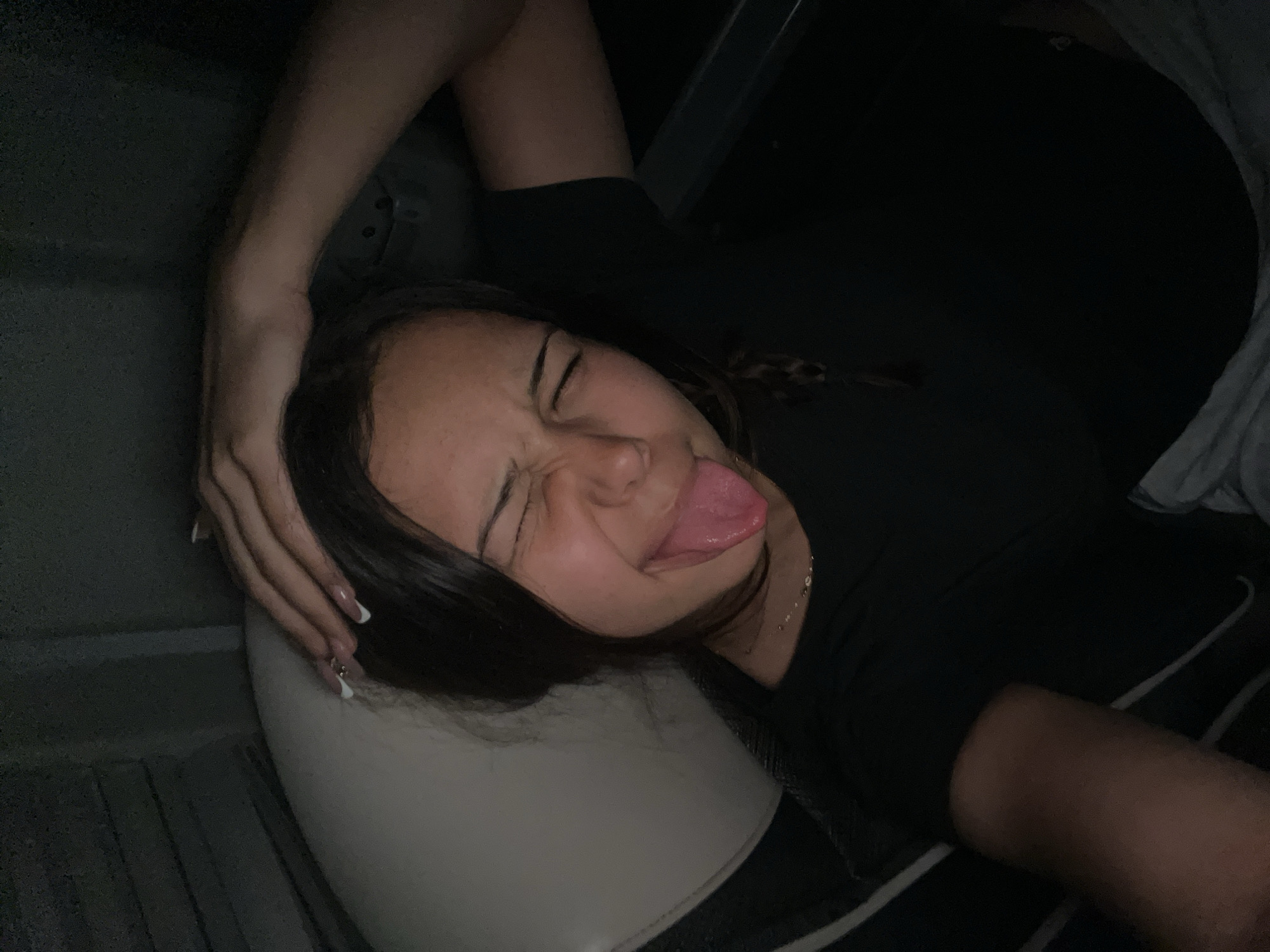 A young woman is lying down, possibly in a car seat, with her head resting on a light-colored headrest. She is sticking her tongue out and has her eyes closed in a grimace. Her right arm is bent and positioned behind her head. She is wearing a dark-colored shirt and a thin necklace. The environment is dimly lit. The city is Unknown, Unknown.