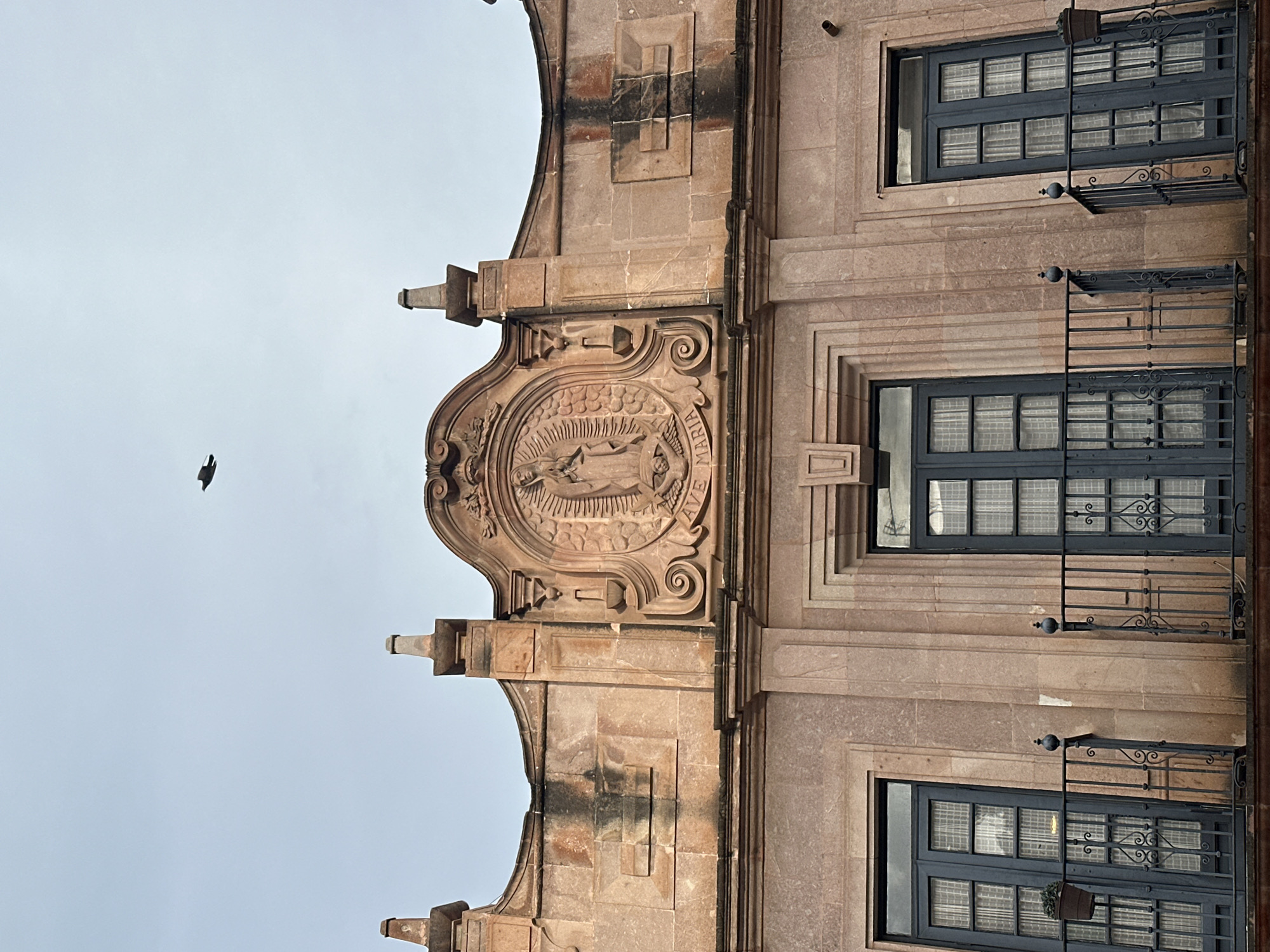 The image shows a view of the façade of a building in Zacatecas City, Mexico, under a cloudy sky. A single bird is flying in the upper center of the image. The building features pinkish-orange stone construction with ornate architectural details. Centered on the upper part of the façade is a bas-relief carving depicting the Virgin Mary, with the inscription "AVE MARIA" below it. Flanking this carving are decorative pilasters and finials. Below the ornate upper section are three windows, each with dark blue double doors and glass panes covered by sheer white curtains. Each window has a wrought-iron balcony with decorative scrollwork. A small potted plant is visible on the balcony to the left. The building exhibits signs of age and weathering, with some darker streaks on the stone.