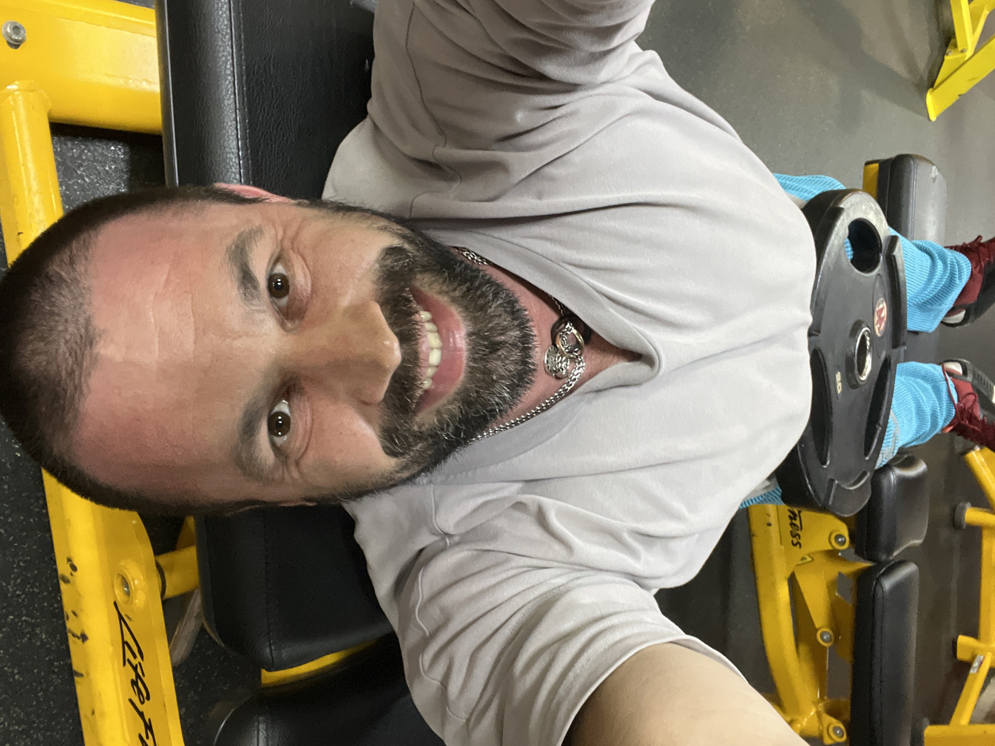 The image shows a first-person perspective of a man lying on a weight bench in a gym. He is wearing a light gray long-sleeved shirt and blue athletic pants. Red and black athletic shoes are visible on his feet. A weight plate rests on his lower body. The gym equipment is yellow and black, with "Life Fit" branding visible on a piece of equipment. The man has a beard and is wearing a silver chain necklace. The floor appears to be dark rubberized matting. The location in Yoshkar-Ola, Russia, is not specified or discernible from the image.