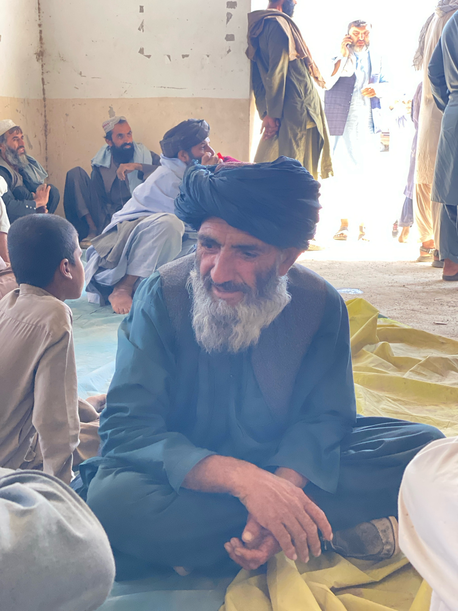 The image shows a group of men sitting on the floor inside a building. The room appears to be simple, with bare walls and a dirt floor. The men are dressed in traditional Afghan clothing, including turbans and long shirts. Some of the men are looking at the camera, while others are looking away. The man in the foreground is wearing a dark blue turban and a matching shirt, and he has a long white beard. He is sitting with his legs crossed, and his hands are clasped in front of him. A young boy in a light brown shirt is visible to the left of the man in the foreground. In the background, other men are visible, some of whom appear to be talking on cell phones or engaged in conversation. One man, on the right side of the image, is wearing a light blue shalwar kameez and a dark vest. The overall impression is one of a gathering or meeting taking place in a communal setting. The environment suggests a rural or semi-rural location, consistent with the city of Pasab, Afghanistan.