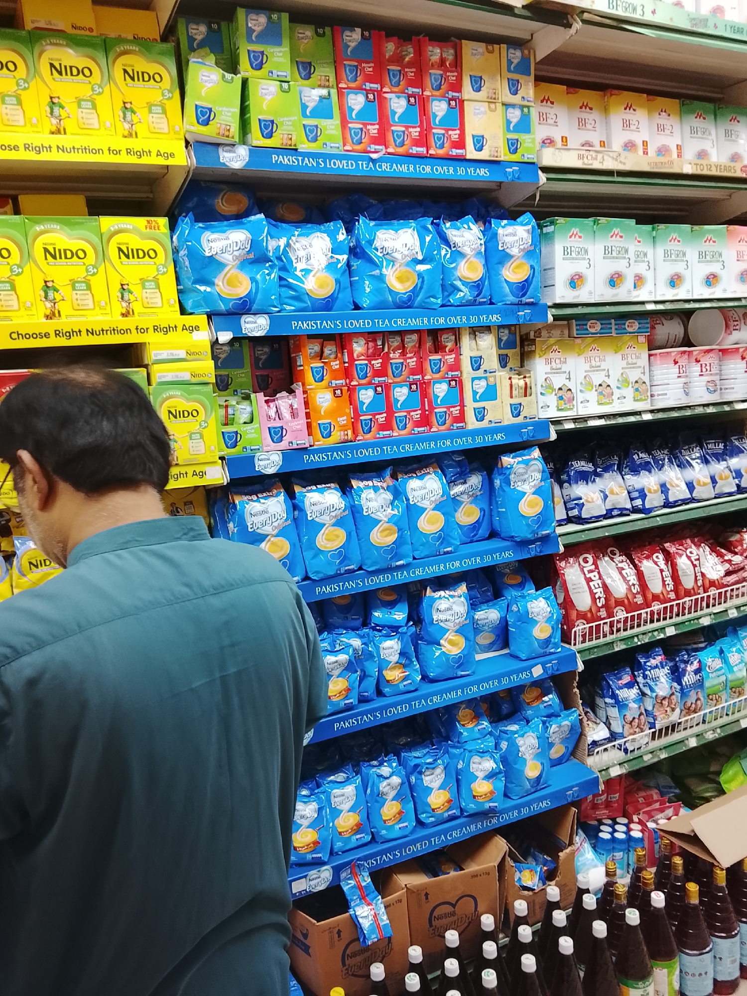 An indoor scene shows a person standing in a supermarket aisle in Gulgasht Colony, Pakistan. The person has short, dark hair and wears a dark teal shirt. The aisle shelves are stocked with various products. On the top shelves, there are boxes of Nido milk powder, tea creamer packets in green, red and orange packaging and infant formula boxes. Beneath those are several shelves filled with blue packets of Nestle EveryDay tea creamer, marked with "Pakistan's Loved Tea Creamer for over 30 Years." Some shelves also hold boxes of BF Chill milk product, Olper's products and other packaged milk powder brands. Brown bottles with white caps sit at the bottom of the shelf.