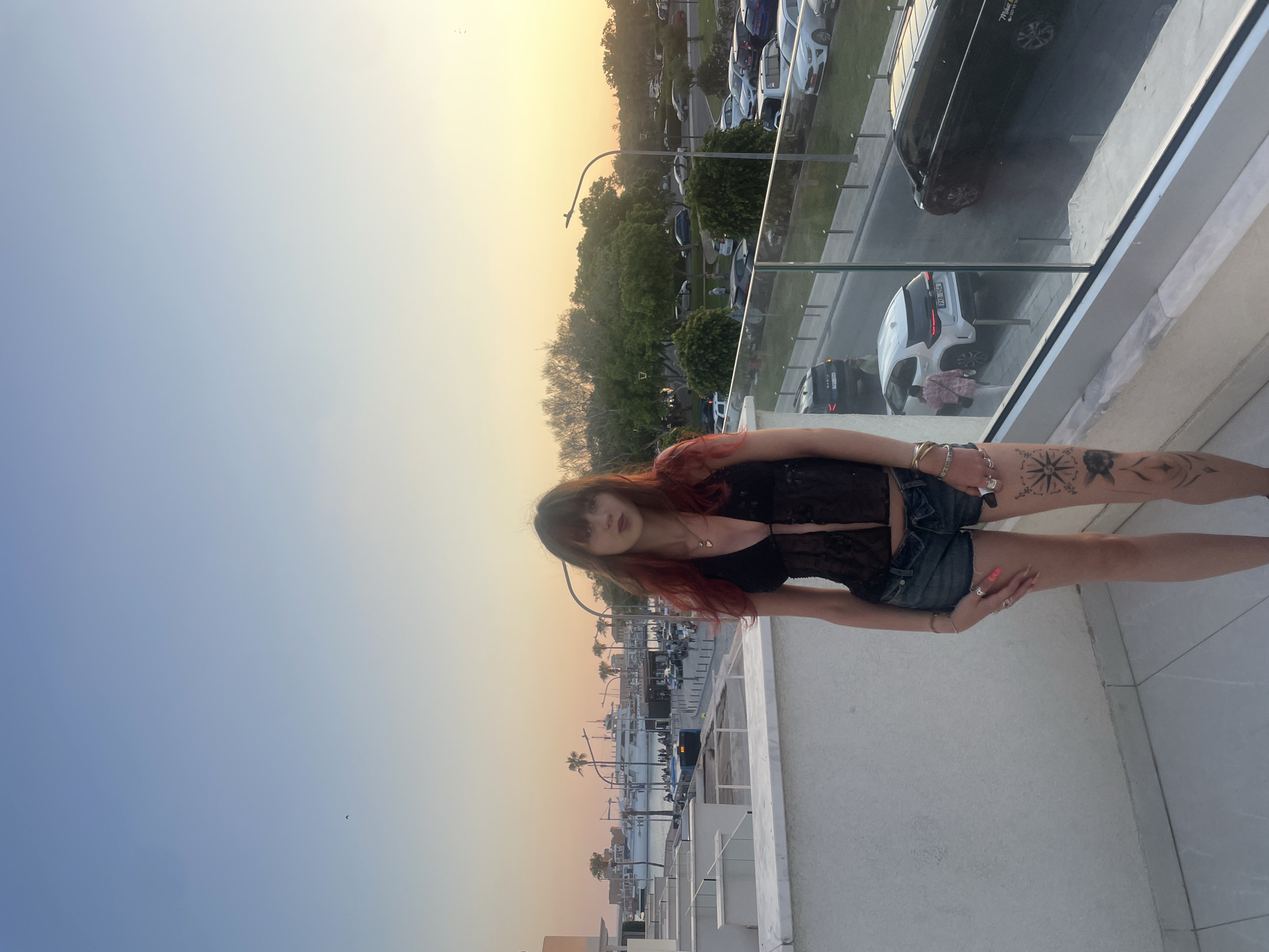 The image captures a young woman standing on a balcony overlooking a waterfront area. The setting is the city of Paphos Municipality, Cyprus, identifiable by the distinctive harbor architecture and palm trees. The woman, with her red hair and a black lace top paired with denim shorts, stands in the foreground. She has tattoos on her legs. Behind her, a harbor with yachts and boats is visible, alongside a street with parked cars and passing vehicles. The time of day is dusk, with the sky exhibiting a gradient of blue and orange hues as the sun sets. The overall atmosphere is relaxed and scenic, suggesting a vacation or leisure setting.