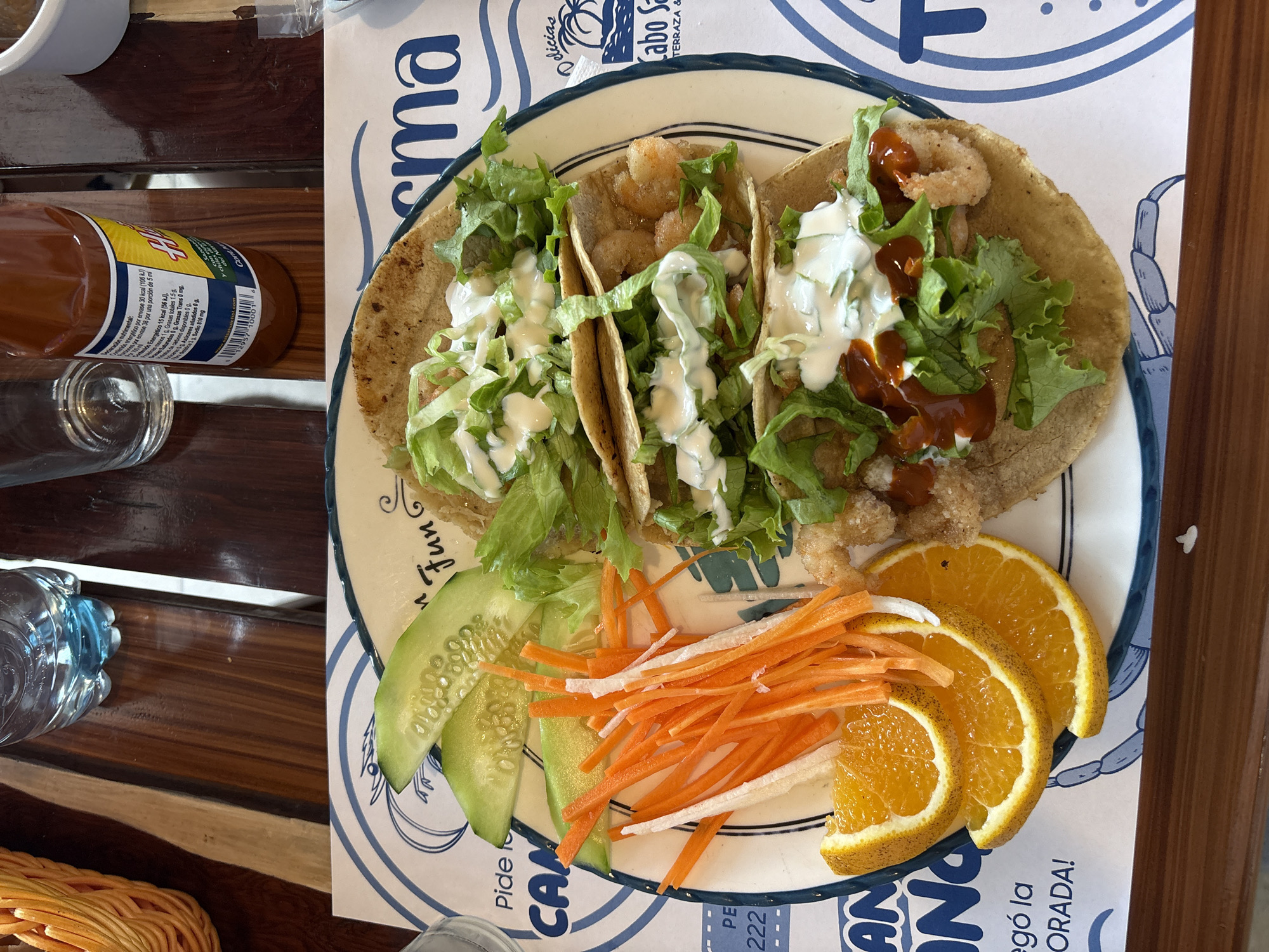The image shows a plate of three shrimp tacos topped with lettuce and a creamy sauce, along with sides of cucumber slices, shredded carrots and white strips, and orange slices. A bottle of Cholula hot sauce and a water bottle are nearby on a wooden table. The plate rests on a paper placemat advertising "Cabo San Lucas Terraza & Fun" with a crab image. The scene is likely in a restaurant located in the city of San Bernardino Tlaxcalancingo, Mexico.