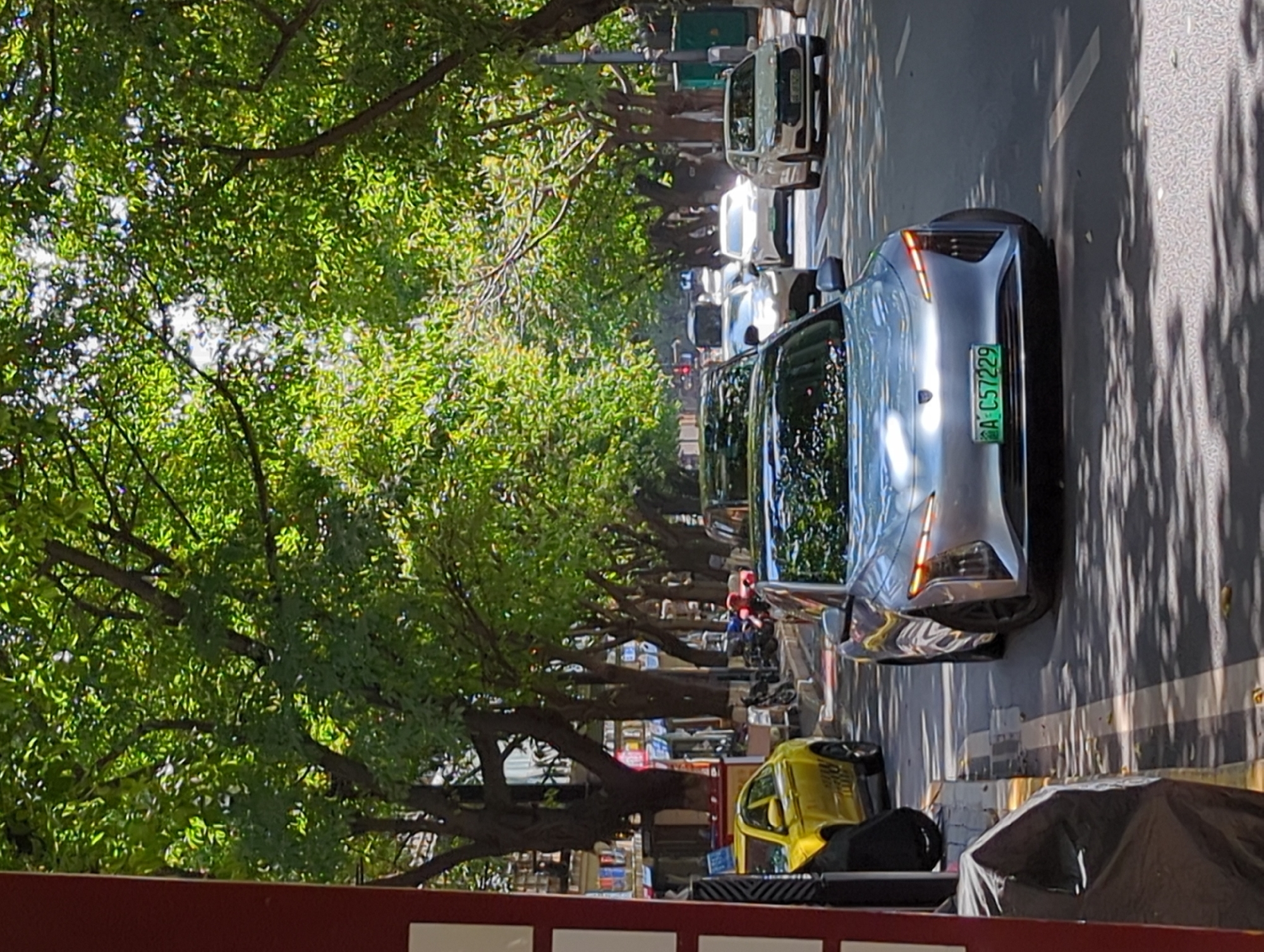 In Yuzhong District, China, a silver electric car with license plate AC57229 is driving on a tree-lined street. The car's headlights are illuminated, and the license plate is a bright green. Several other cars are also visible on the road, including a yellow vehicle parked on the side. Buildings and shops line the street, and the trees overhead cast dappled shadows on the asphalt. The sun is shining, creating a bright and airy atmosphere.