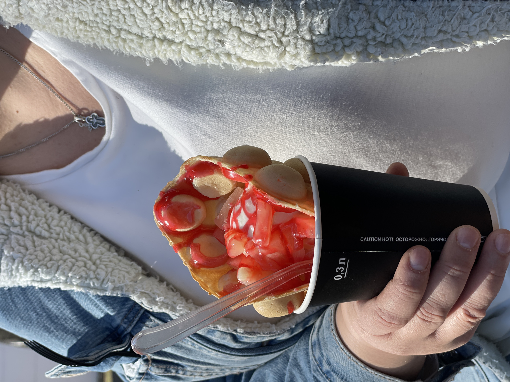 The image shows a person holding a black cup filled with a bubble waffle and what appears to be a strawberry-flavored topping and ice cream. The person is wearing a white shirt, a white sherpa-lined jacket, and a blue denim jacket. A silver necklace is visible around the person's neck. The text "CAUTION HOT! ОСТОРОЖНО: ГОРЯЧО" is printed on the cup, along with "0,3 л". A clear plastic spoon is inside the waffle cup. The setting appears to be outdoors. The location is Unknown, Unknown.