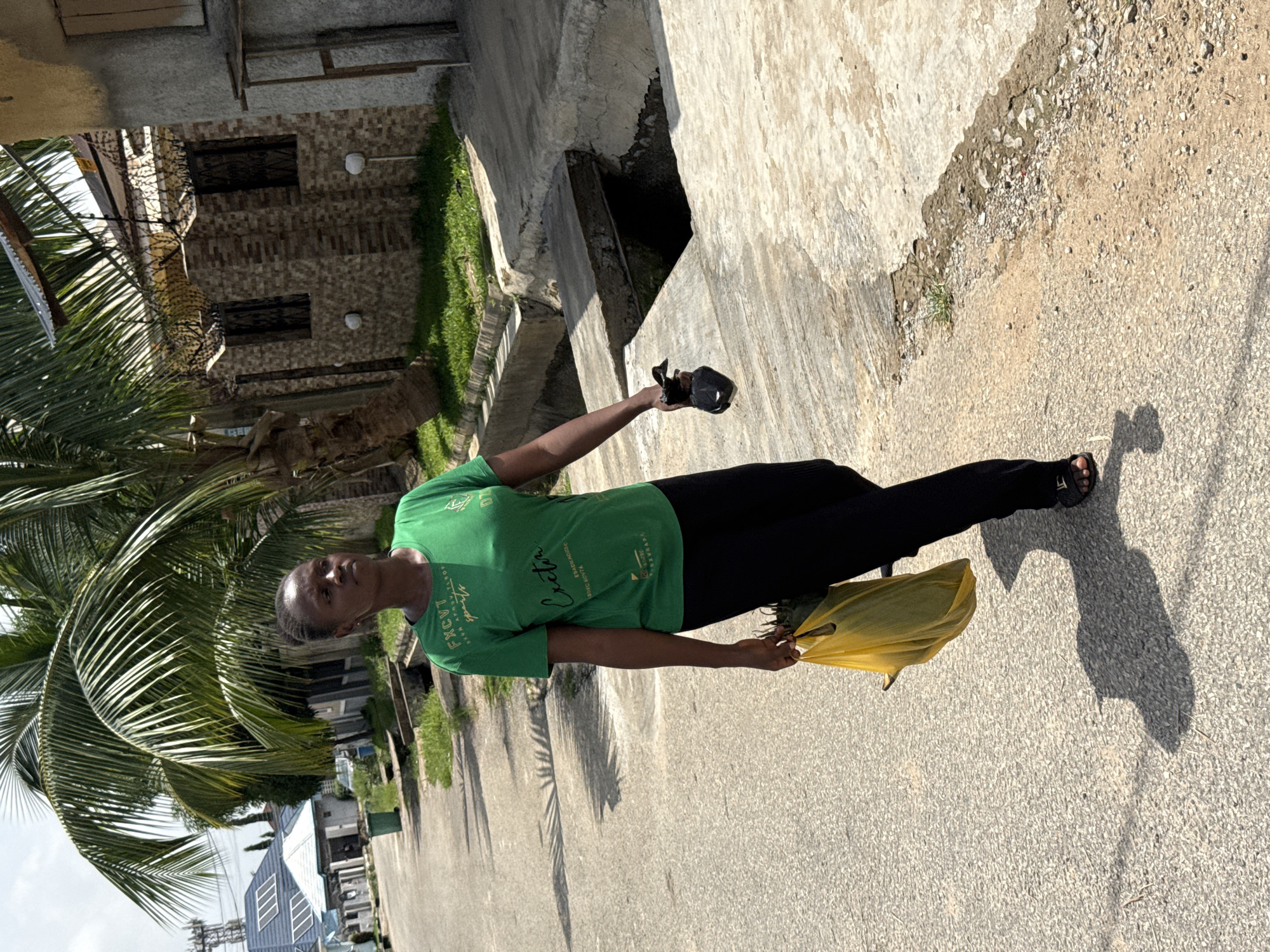 A middle-aged woman is standing on a gray concrete street in Abuja, Nigeria. She is wearing a green t-shirt and black pants, and she is holding a yellow plastic bag in her left hand and a black object in her right. To her left is a large palm tree. In the background, there is a building with a stone facade and barred windows. A concrete structure with a drain opening is adjacent to the street. The location is a residential or suburban area.