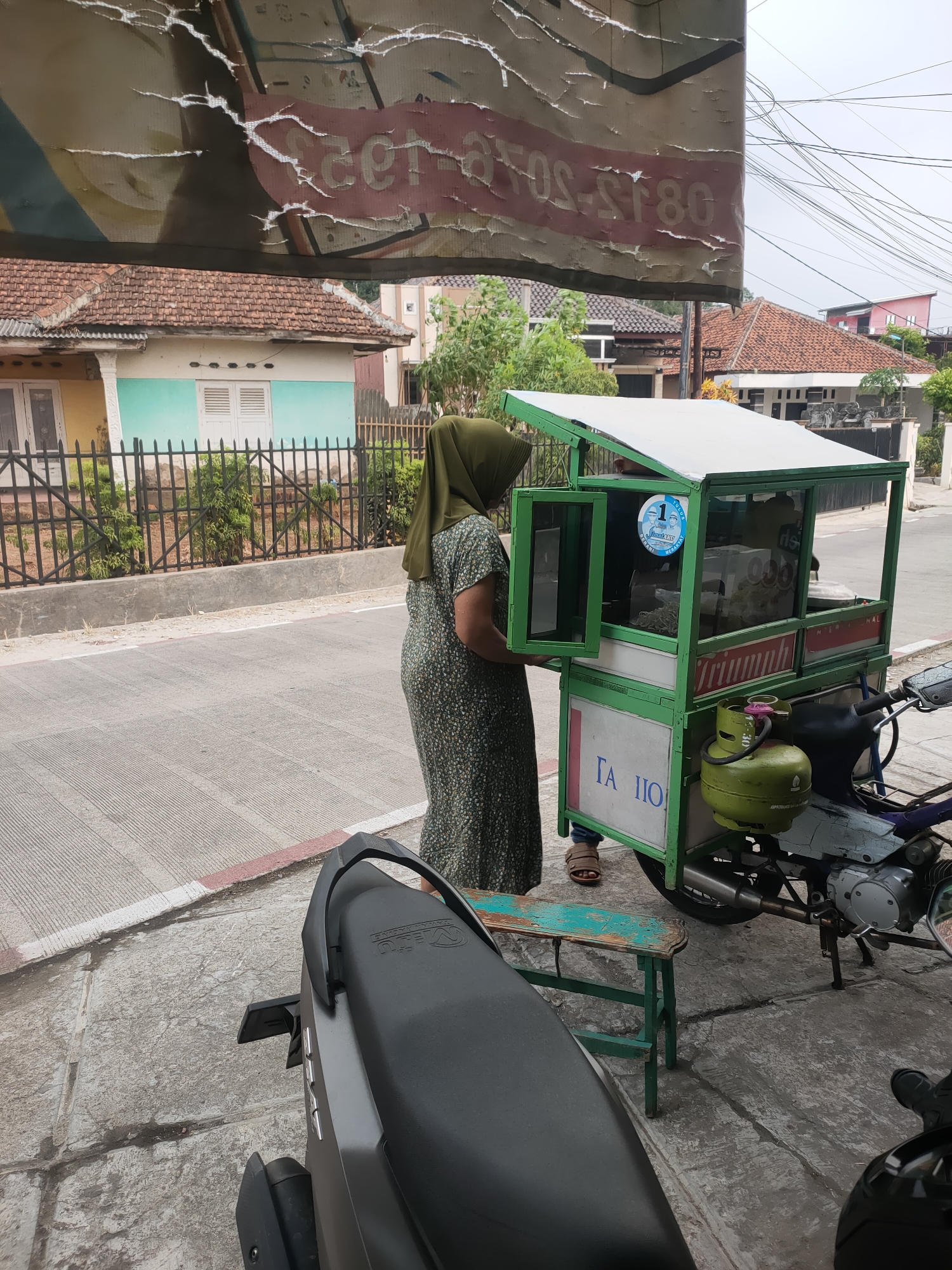 In Subang, Indonesia, a woman wearing a green hijab and a patterned dress stands beside a mobile food cart. The cart, painted green and white, is mounted on a motorcycle and has a display case showcasing food items. A green gas cylinder is attached to the side of the cart. A small, weathered green stool is placed on the ground in front of the cart. In the background, houses with tiled roofs are visible, along with trees and a cloudy sky. A torn banner hangs overhead, partially obscuring the scene. The woman appears to be interacting with the food cart, possibly a vendor or a customer.