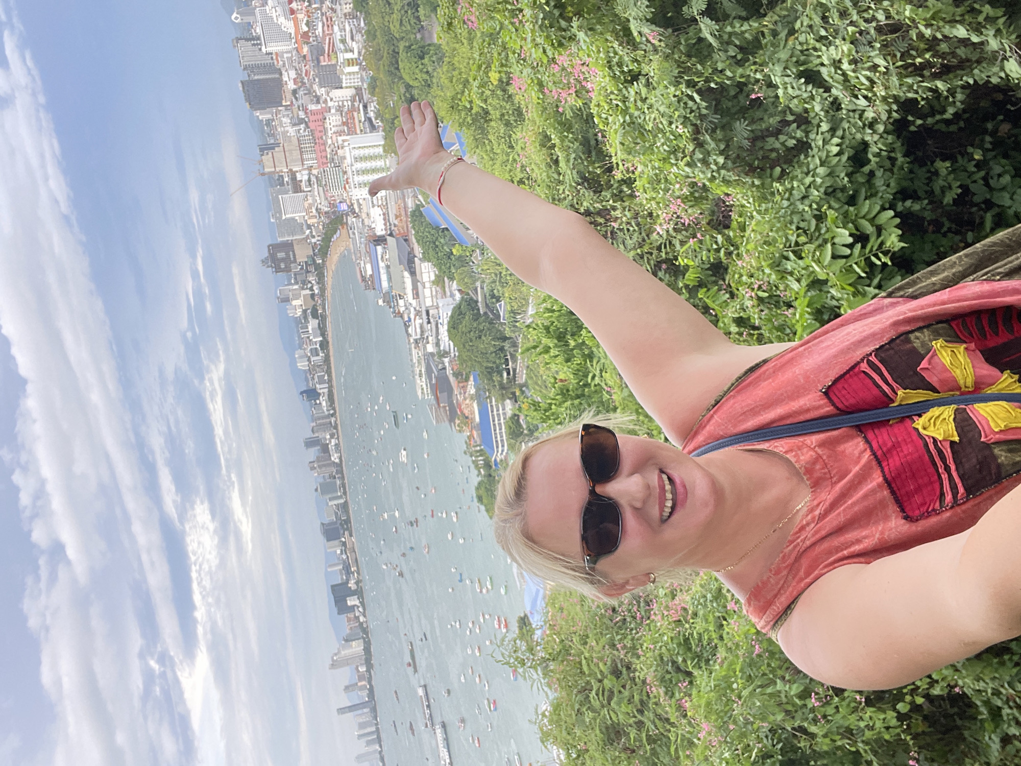 In Pattaya City, Thailand, a woman with blonde hair and sunglasses smiles at the camera. She is wearing a coral-colored tank top with a decorative patch and a blue strap is visible over her shoulder. Her right arm is raised and extended towards the viewer. The background features a panoramic view of Pattaya's coastline, with numerous boats scattered across the turquoise water. Beyond the water, a densely populated city skyline rises, characterized by a multitude of buildings of varying heights. Lush green foliage and flowering plants in the foreground frame the woman and the cityscape. The sky above is partially clouded, with a mix of blue sky and white clouds.