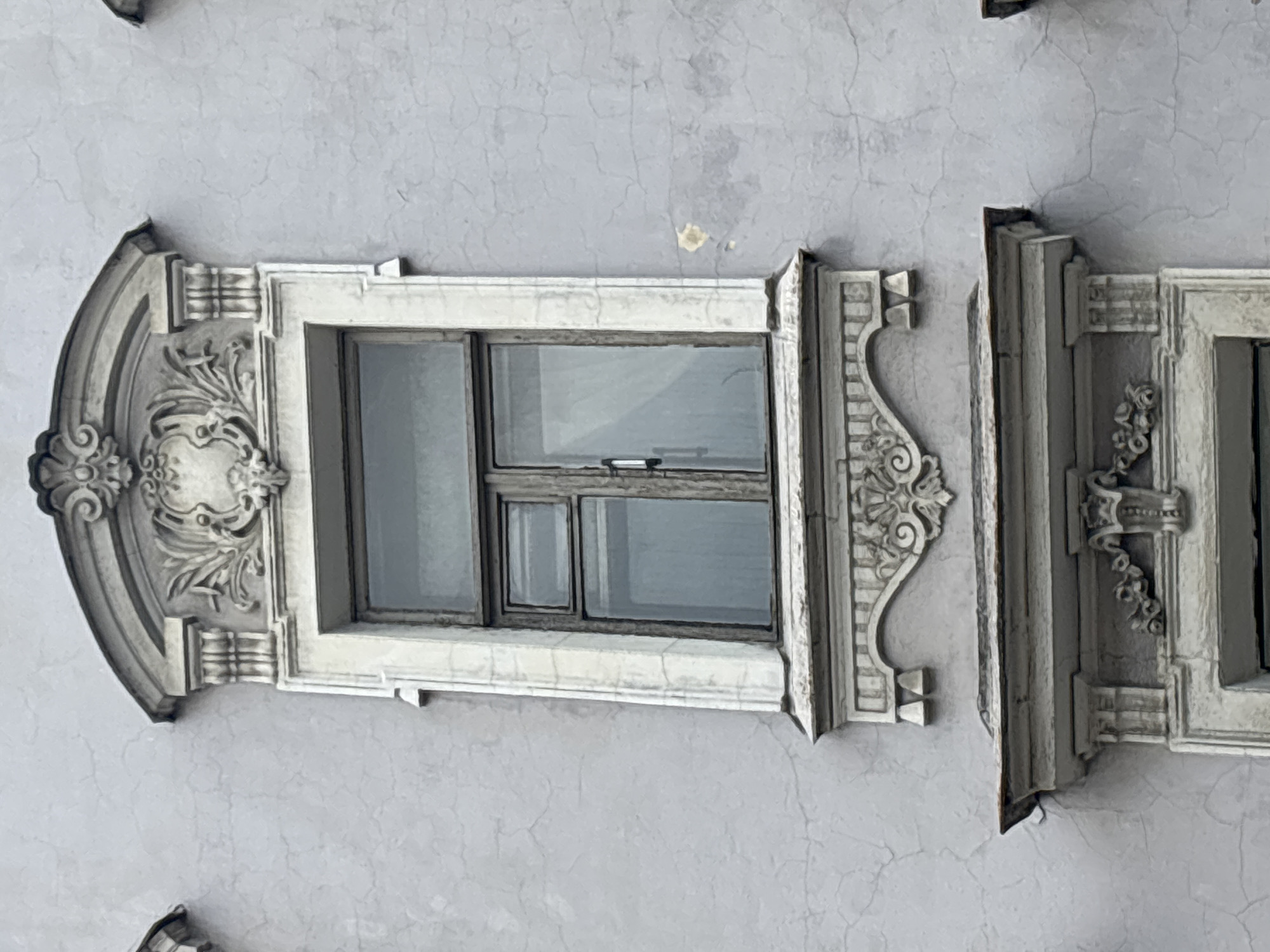 A close-up, eye-level, medium shot captures a segment of a weathered, light grey building facade in Saint Petersburg, Russia, featuring an ornate window. The window itself is framed by white decorative plasterwork, characterized by volutes, floral motifs, and what appear to be acanthus leaves. The upper portion of the window frame is crowned by an elaborate pediment with a central cartouche and flanking decorative elements. Below the window, a projecting ledge or sill with similar decorative plasterwork is visible, extending outwards from the facade. The window panes are divided into multiple sections, with the frames appearing to be made of dark wood. The overall impression is one of aged architectural grandeur, with some visible cracking and imperfections in the building's plaster. The lighting is even, suggesting an overcast day.