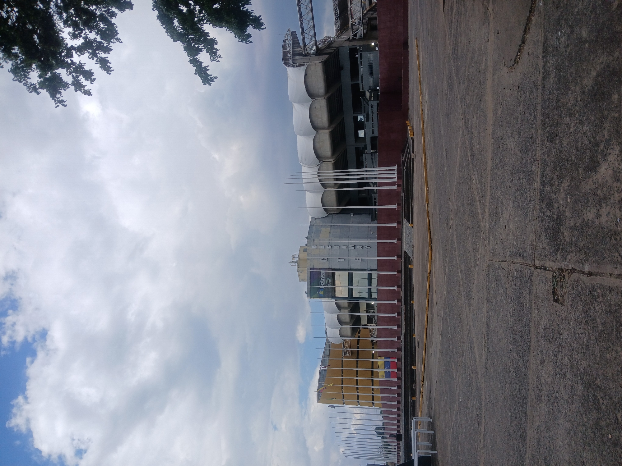 The image shows a stadium in Ciudad Guayana, Venezuela, viewed from street level. The stadium features architectural elements including a covered seating area with arched supports and a taller section with a multi-level structure, banners, and signage. A row of flagpoles lines the perimeter of the stadium. The foreground is a concrete surface, and the sky above is partly cloudy. Vegetation is seen in the upper left corner.