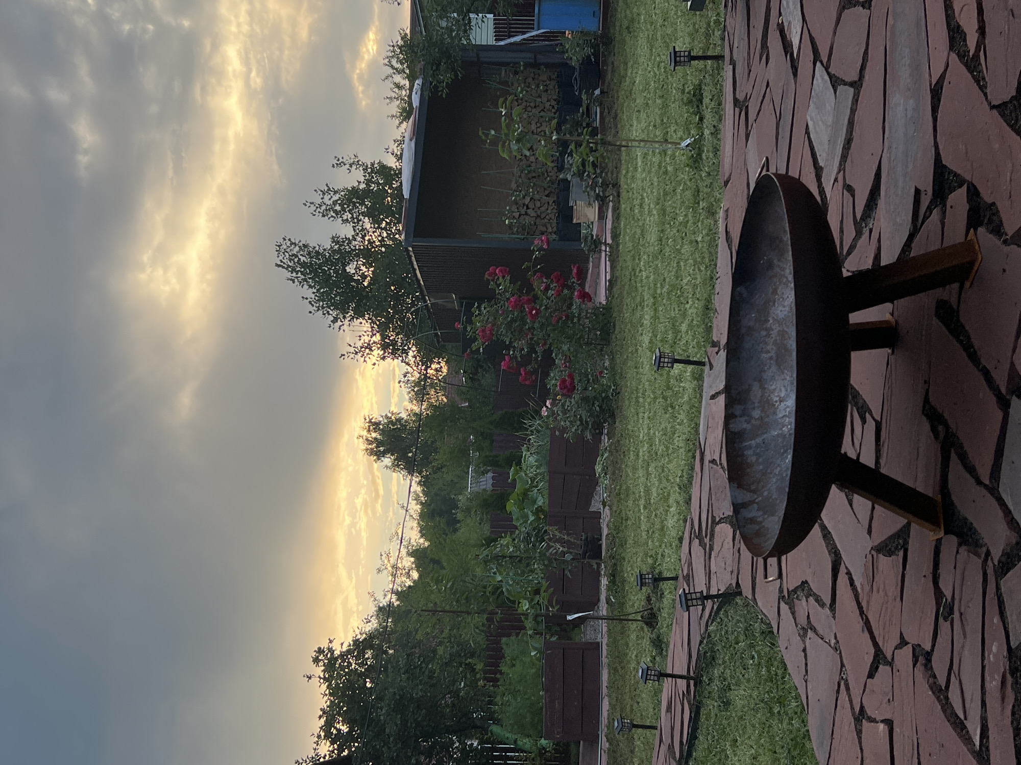 The image depicts a backyard scene at dusk, likely in Luch, Russia, given the style of the wooden raised garden beds and the overall rustic aesthetic. In the foreground, a metal fire pit with three wooden legs rests on a patio paved with irregularly shaped red flagstones. A winding pathway of similar flagstones leads into the yard, bordered by small solar-powered garden lights.
The yard features a neatly mown lawn and several garden beds filled with lush greenery, possibly vegetables. A prominent rose bush with vibrant red blooms stands to the left of a utilitarian metal structure, possibly a woodshed or storage unit, which is filled with neatly stacked firewood. Beyond the immediate garden area, mature trees and foliage create a natural backdrop, with a wooden fence visible in the distance.
The sky is dramatic, with heavy grey clouds tinged with the warm hues of sunset, suggesting either the end of a stormy day or an approaching storm. The lighting is dim, casting long shadows and creating a tranquil, somewhat melancholic atmosphere. There are no people or animals visible in the scene.