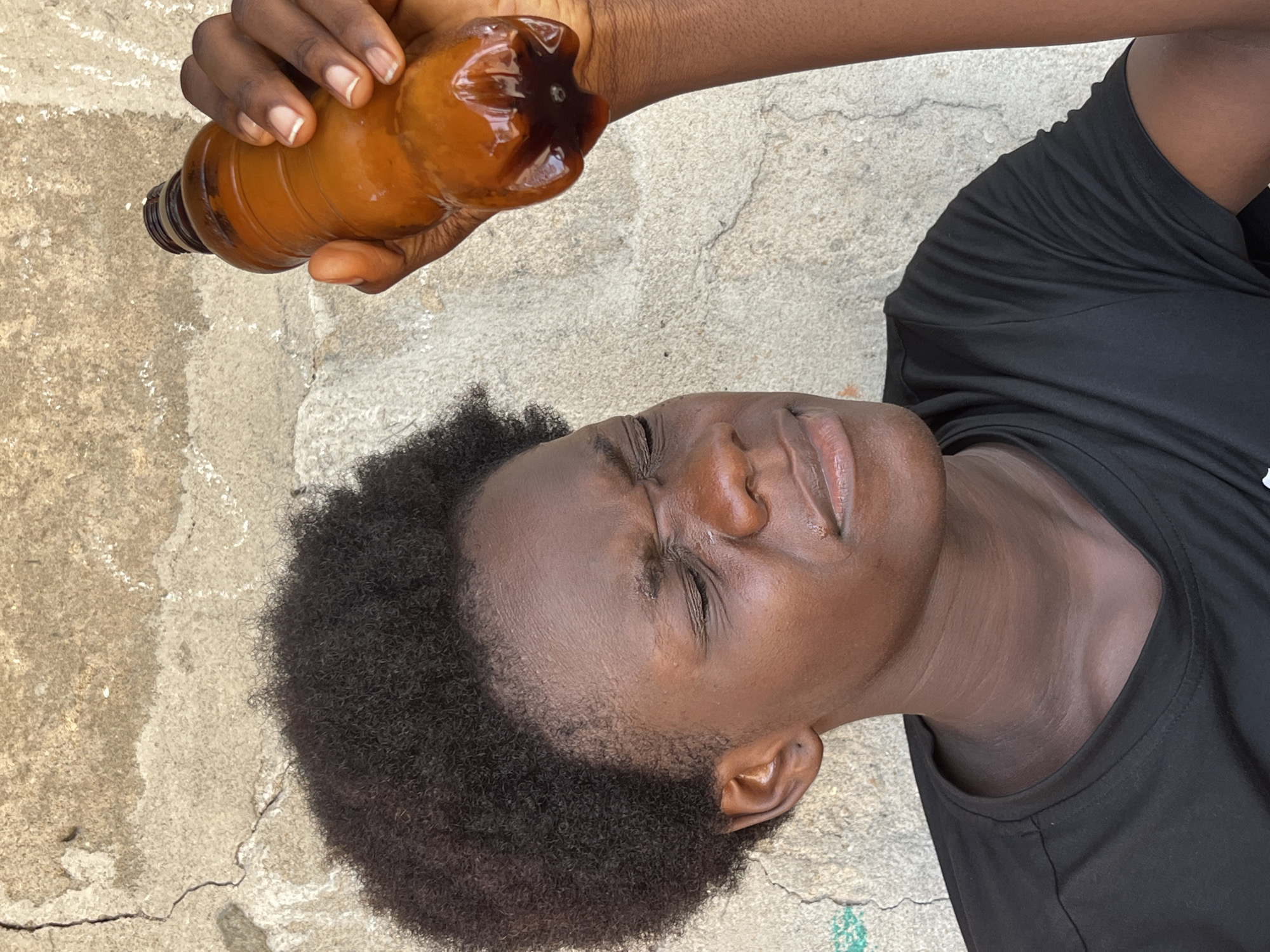 A dark-skinned person is holding a plastic bottle of dark liquid over their face. The person has short, dark curly hair, their eyes are closed, and they are wearing a black t-shirt. They are outside on a concrete surface, which appears to be cracked and discolored in sections. The location is Badagry, Nigeria.