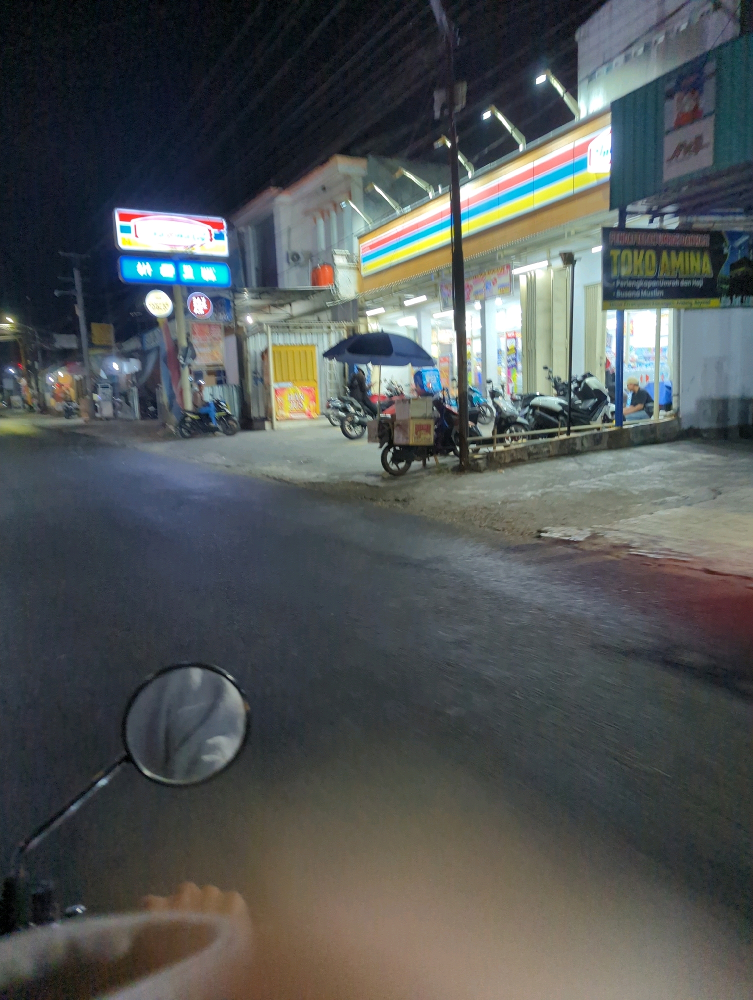 The image depicts a street scene at night in Kacangan, Indonesia. In the foreground, the handlebars and a rearview mirror of a motorcycle are visible, suggesting the photo was taken from the rider's perspective. The road surface is dark asphalt. On the right side of the road, a convenience store with a brightly lit facade is prominent. The store's signage includes "Indomaret" in red and white, and another sign reads "TOKO AMINA" in yellow lettering with smaller text below. Several motorcycles are parked in front of the store, and a person is seated on a bench outside. An umbrella shades a small food cart with boxes. In the background, other buildings and streetlights are dimly illuminated against the night sky.