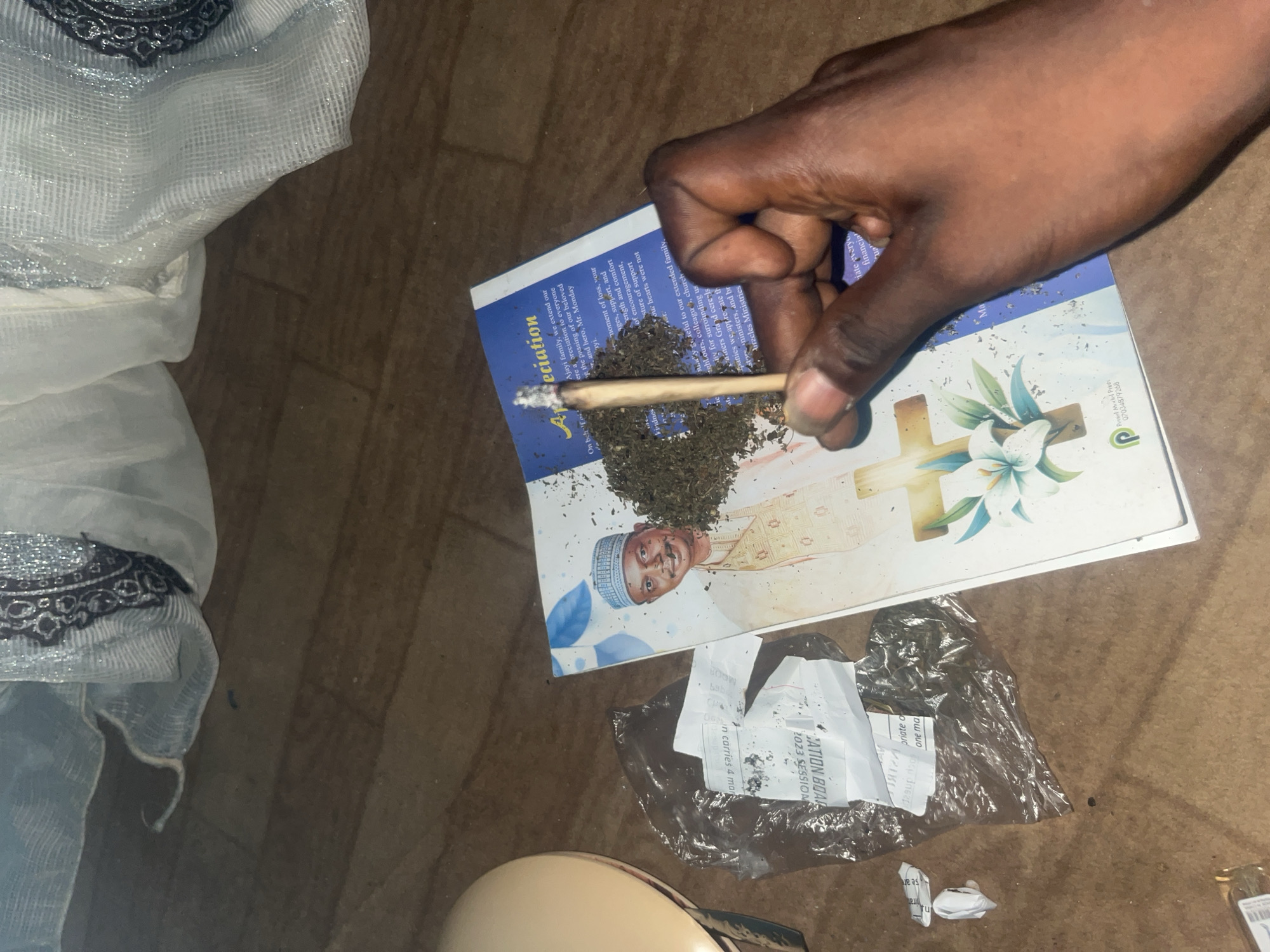 A hand holds a lit marijuana cigarette above a booklet that has a picture of a man in traditional attire. Scattered dried leaves are also present on the booklet and surrounding area. A plastic bag containing what appears to be more dried leaves and paper is to the left of the booklet. The scene is set on a wooden floor, with sheer white curtains in the background. The image does not provide any information about the location in Gura Top, Nigeria.