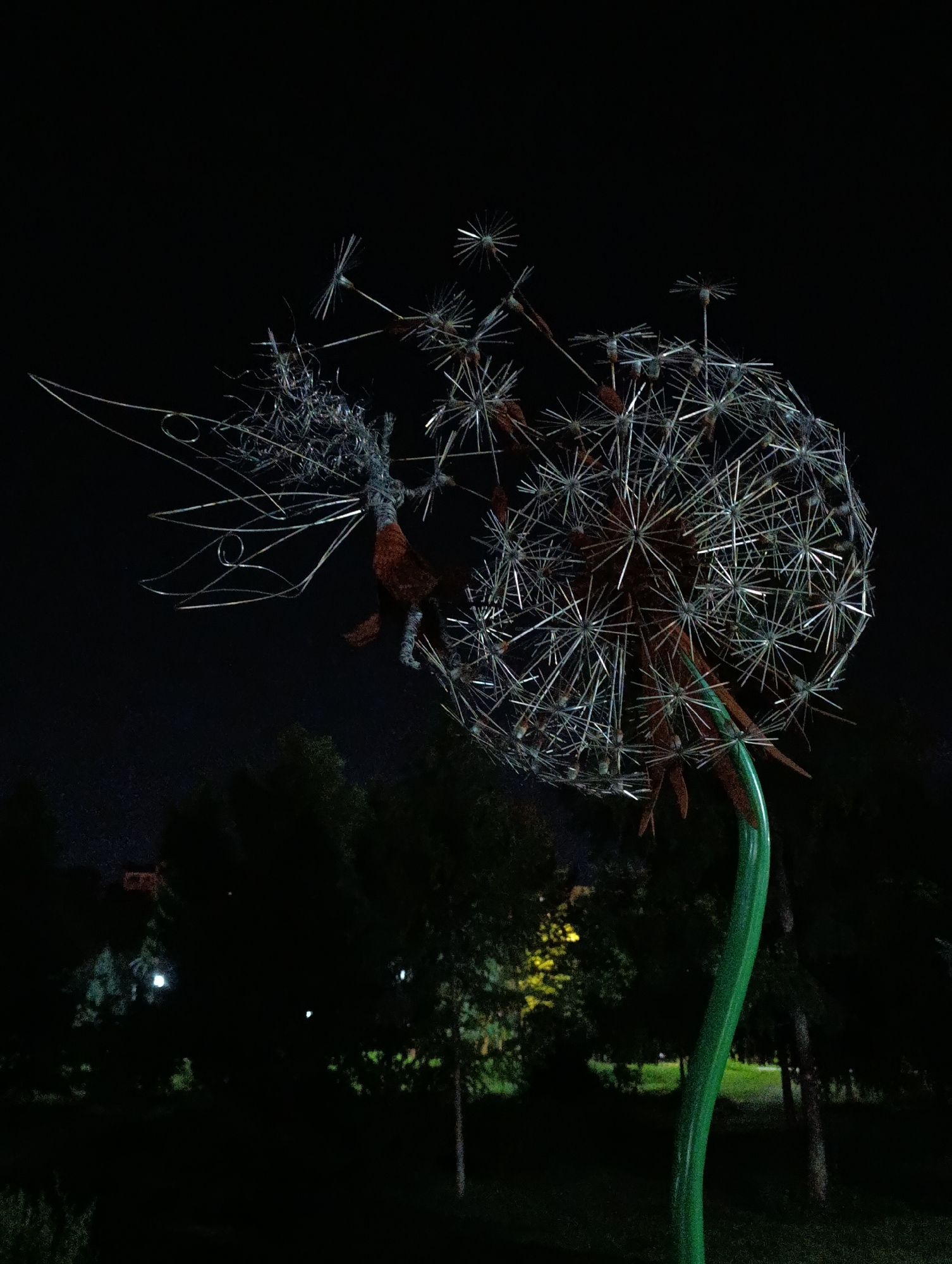This is a nighttime photograph of a large metal sculpture of a dandelion seed head. The sculpture is lit from below, highlighting its intricate wire frame and the reddish-brown interior of the seed head. The stem of the dandelion is a vibrant green and curves upwards. The background is dark, with the silhouettes of trees and some faint lights visible in the distance, suggesting an outdoor park or public space. The photograph appears to have been taken in Abakan, Russia.