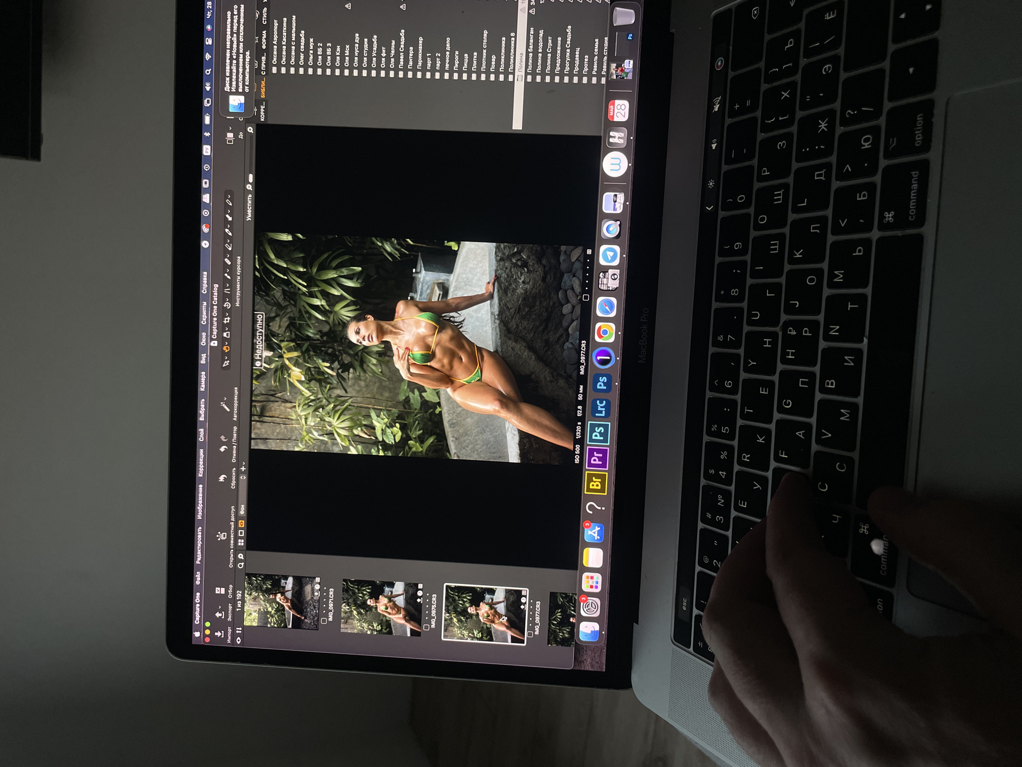 A person is using a MacBook Pro to edit a photo in Capture One, a photo editing software. The photo being edited features a woman in a green bikini posing outdoors. The MacBook is a Russian keyboard model. The location is identified by the file names and folders on the screen, with Russian names such as "Oksana Kasatkina," "Olga Svadba," "Polina," and "Ravil's studio." Based on these, the image editing is being done in Kazan, Russia.