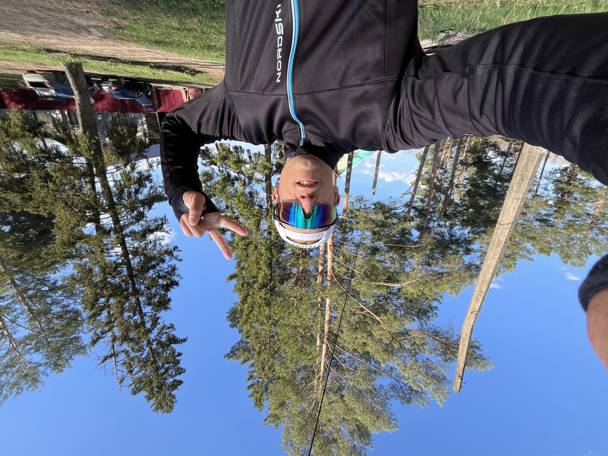 A man is holding his phone while standing outside during the day and taking a selfie. He is wearing a black jacket, colorful mirrored sunglasses, and a white headband. He is giving a peace sign with his left hand.
Behind him is a blue sky and tall green trees. The photo is oriented upside down.
Based on the architecture visible in the background, and the jacket, it is likely the location is near Chaikovsky, Russia.
