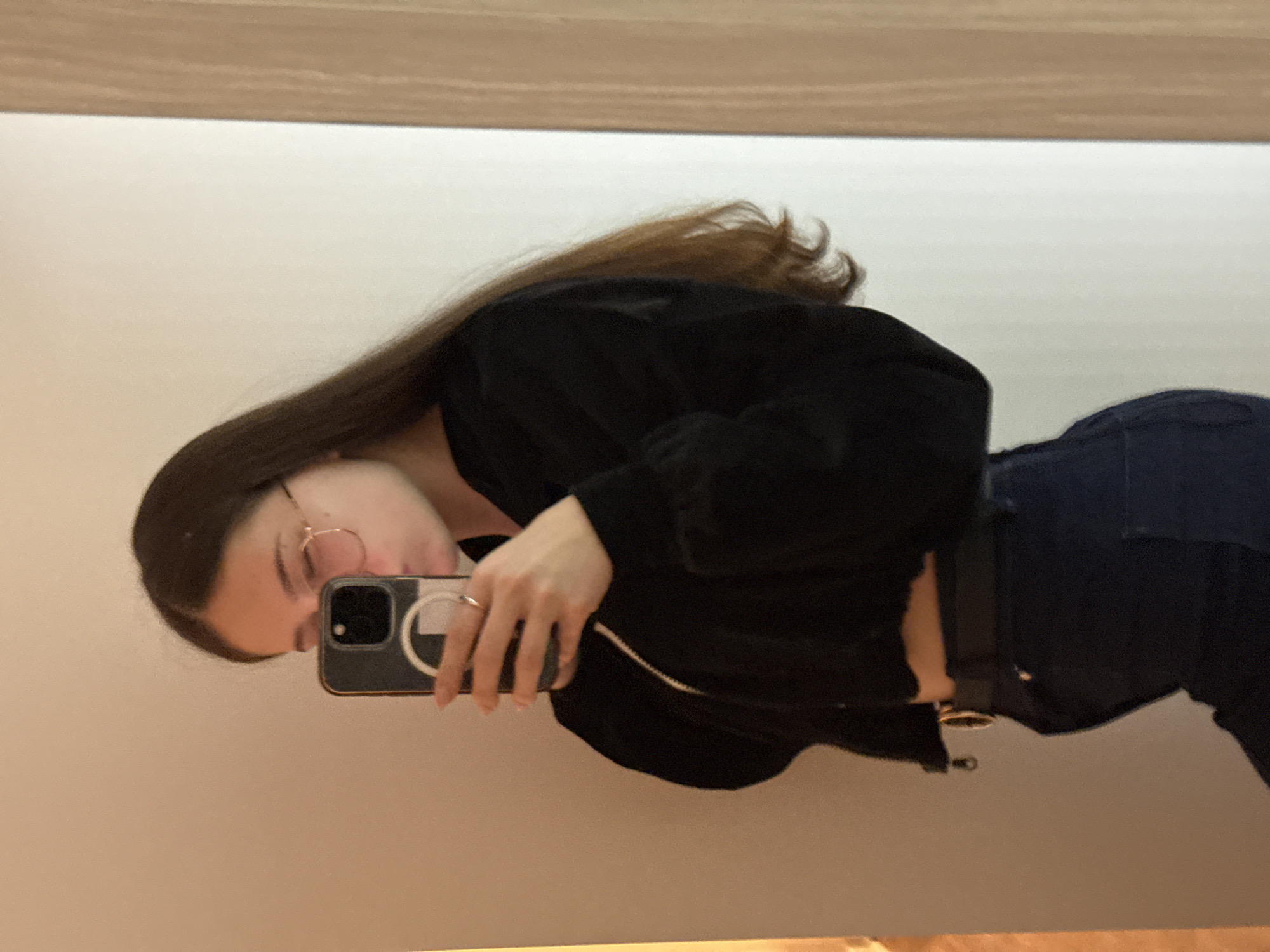 A female individual is photographed in a mirror reflection, visible from the torso up, within an interior setting. She has long, straight dark brown hair extending over her shoulders and back. She wears round, thin-framed eyeglasses. Her right hand holds a dark-colored smartphone, featuring a prominent white circular design on its case, positioned in front of her lower face, with her lips appearing slightly pursed. A silver-colored ring is present on her right index finger. She is dressed in a black cropped zippered jacket, revealing a segment of her midriff, and dark blue trousers secured by a black belt with a gold-colored circular buckle. The background consists of a light beige wall on the left, displaying warmer illumination, transitioning to an off-white section on the right, which is bordered by a vertical light-colored wooden panel. The scene is situated in the city of Vozdovac Urban Municipality, Serbia.