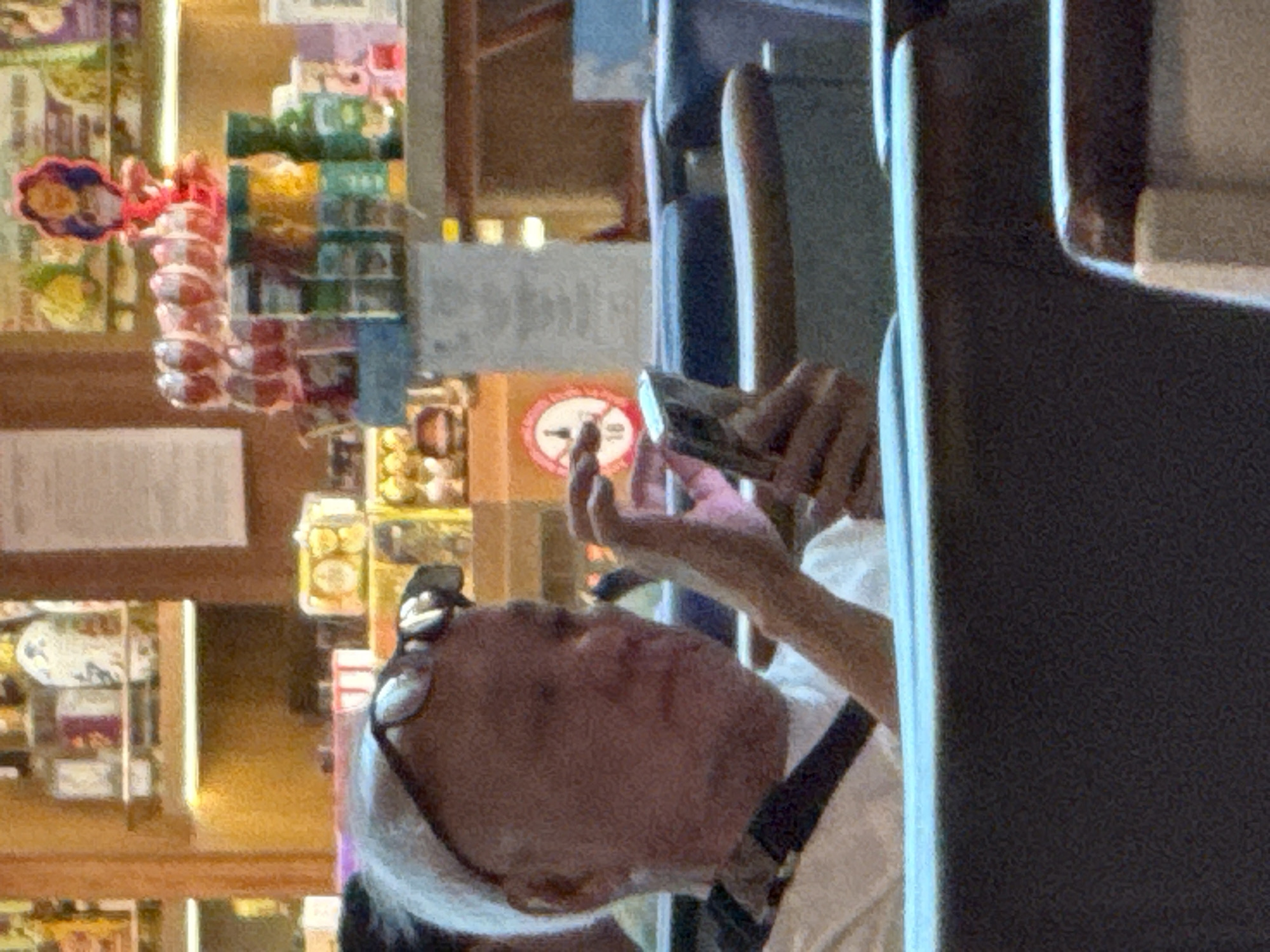 The image shows a person with white hair wearing glasses, sitting and looking at a smartphone. The person is wearing a white shirt and a black strap across their chest. They are holding the phone with their right hand, and their index finger appears to be interacting with the screen. The background suggests a retail environment with shelves stocked with various items, possibly in a duty-free shop or a store in an airport in Bắc Cam Ranh, Vietnam. There are bottles, jars, and other products visible on the shelves. The overall lighting is warm and seems to be artificial, typical of an indoor commercial space.