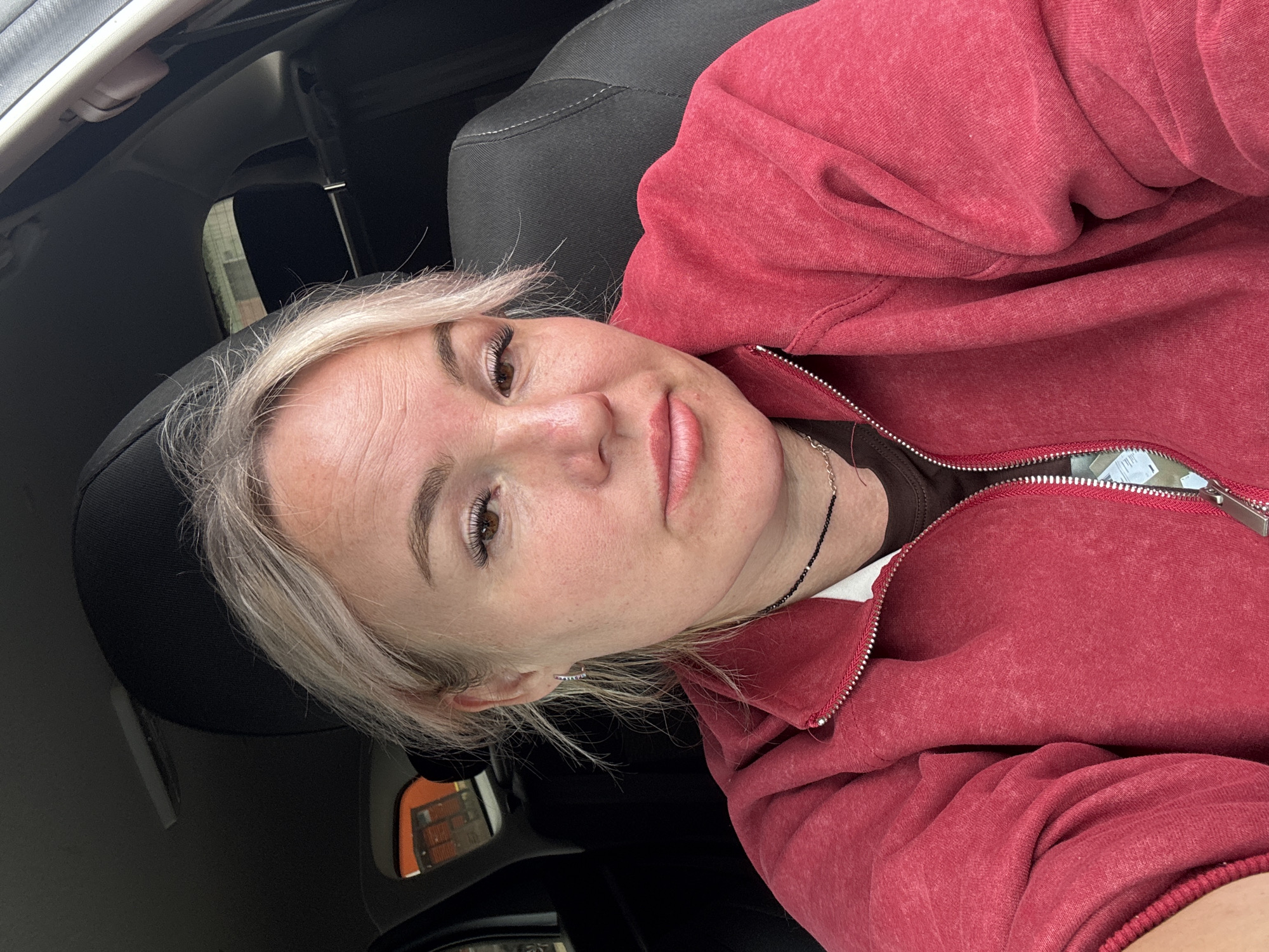 A woman with blonde hair and light skin, wearing a red zip-up hoodie over a dark shirt and a beaded necklace, is sitting in the driver's seat of a car. She is looking towards the camera with her mouth slightly open. The interior of the car, including a black headrest and seat, is visible. Outside the car window, some orange structures and a building can be seen. The image appears to have been taken during the daytime.