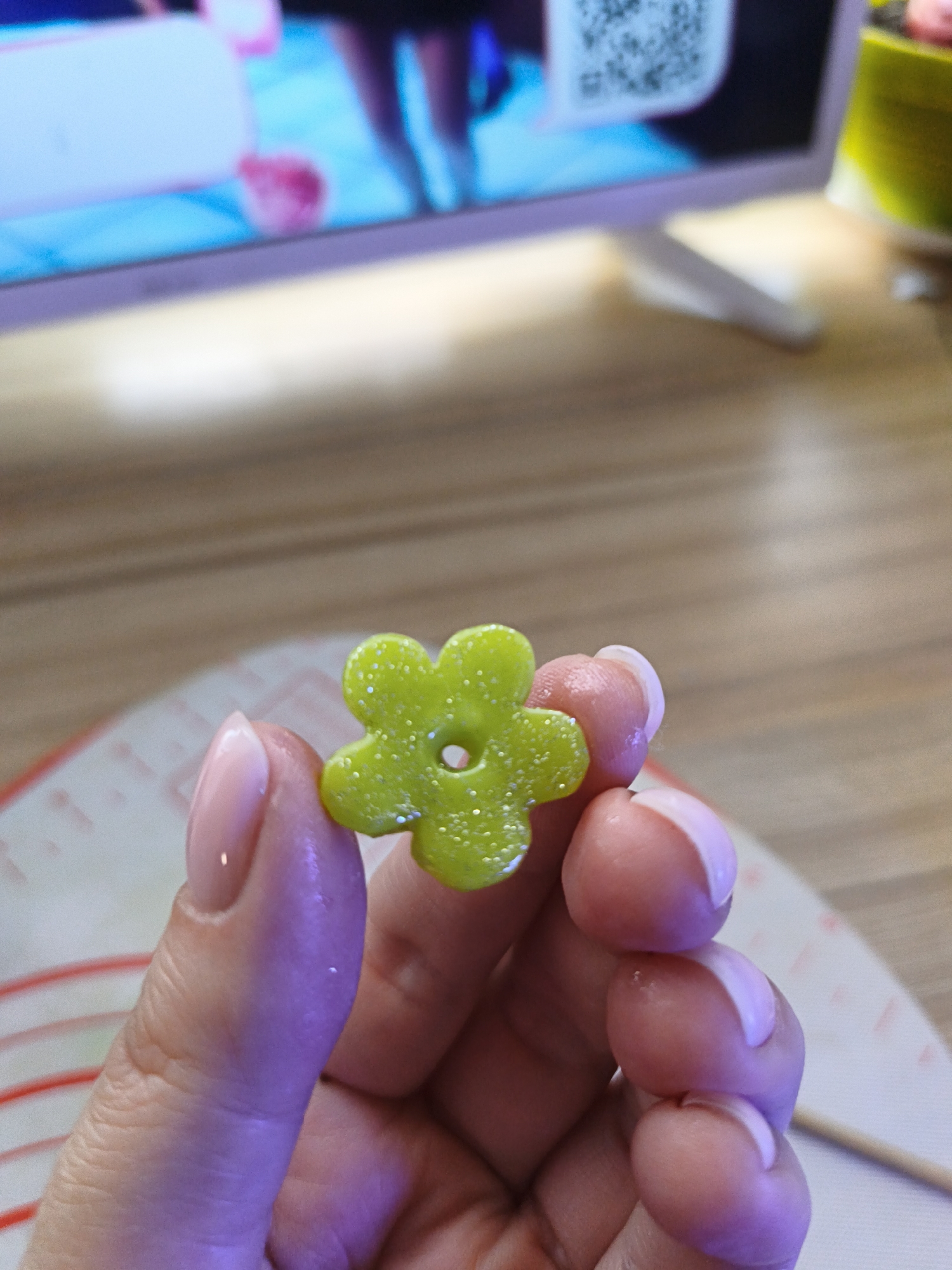 A close-up shot features a person's hand holding a small, vibrant green, flower-shaped object. The object is translucent and appears to be made of resin or a similar glossy material, embedded with tiny, sparkling glitter specks. It has five rounded petals and a small circular hole in its center, suggesting it could be a bead, charm, or decorative piece.
The hand, likely belonging to a female, has neatly manicured nails with a natural pink base and lighter tips, resembling a French manicure. The fingers gently grasp the object, with light reflecting off the skin and nails.
In the blurred background, a screen is visible, displaying bright blue and white imagery. A distinct QR code is present on the right side of the screen, though its content is illegible due to blurriness. Below the screen, a light brown wooden surface with visible grain extends across the frame. Part of a white mat with red horizontal lines, possibly a silicone crafting mat, is visible beneath the hand. Further in the background, a small yellow or lime green object, possibly a container, can be seen. A thin wooden stick or skewer is also partially visible on the mat to the right. The overall lighting suggests an indoor setting, with a mix of ambient and possibly artificial light creating reflections on the shiny surfaces.