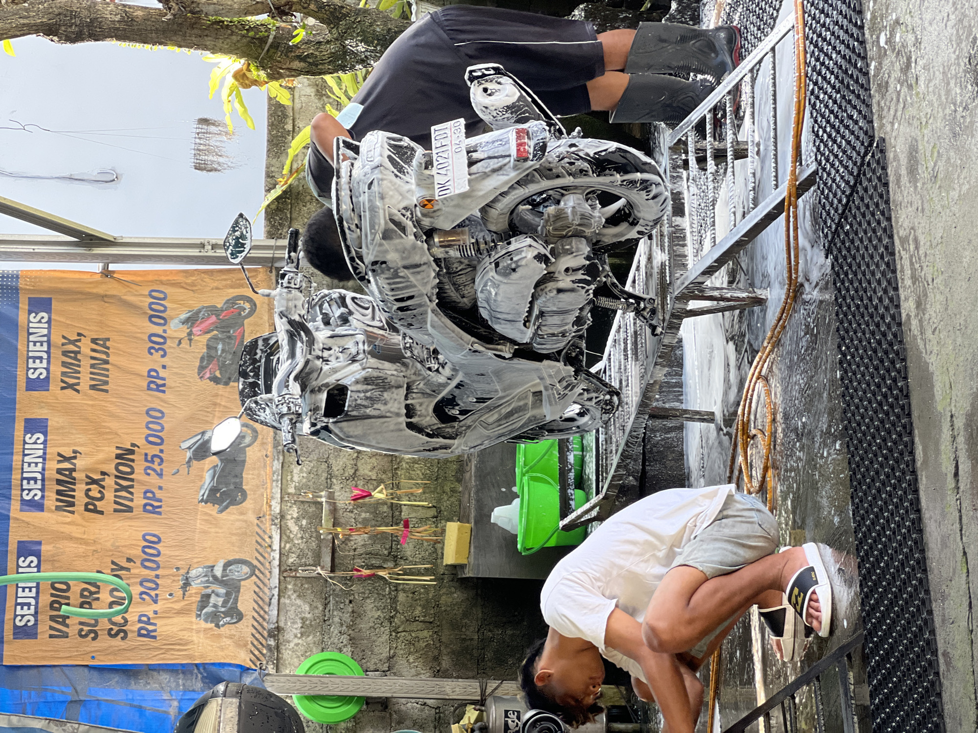 In North Kuta, Indonesia, a motorcycle is being washed at an outdoor service station. A young man in a white t-shirt and light-colored shorts is crouched down on a wet, black textured mat, scrubbing the motorcycle with a brush. He is wearing white and black slide sandals. Another individual, partially visible, stands behind the motorcycle wearing a dark shirt and shorts with black boots, also appearing to be involved in the washing process. The motorcycle, covered in white foam, is positioned on a metal stand. A large banner displaying different types of motorcycles and prices is visible in the background. Green buckets and a yellow sponge are also present, along with hanging decorations and a concrete wall. The scene is outdoors, with a tree to the right and overhead scaffolding suggesting a covered workspace.