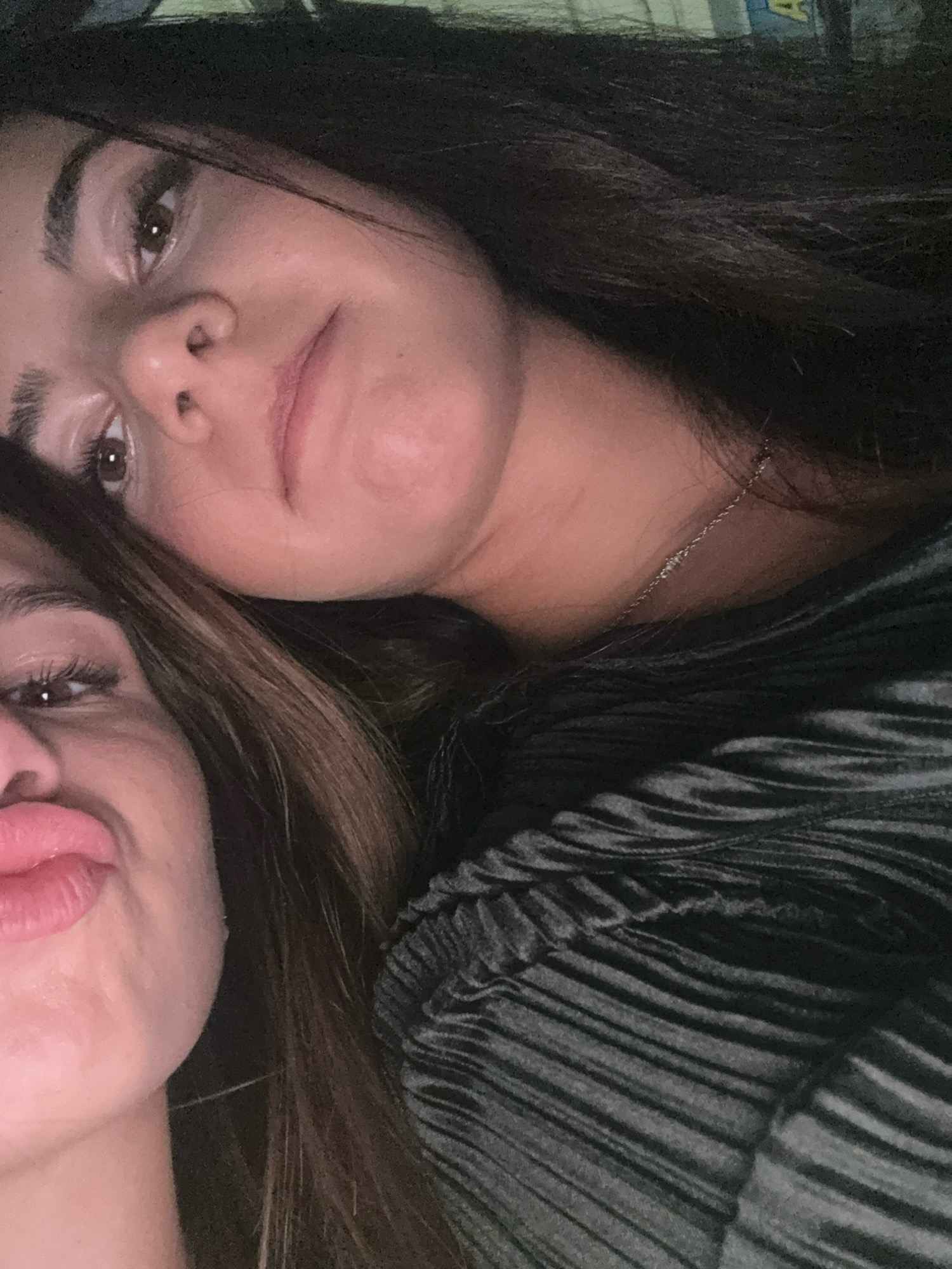 Two women are captured in a close-up, selfie-style photograph, their faces partially obscured and angled towards the camera. One woman's head rests against the other's shoulder, her dark hair flowing around them. She is wearing a dark, possibly velvet, top with a textured, ribbed design. A delicate silver necklace with small bead-like accents is visible around her neck. The other woman's face is also in view, with her lips pursed in a playful pout. The lighting is dim, suggesting an indoor setting, possibly a private residence or a dimly lit establishment. The background is dark and indistinct, offering no specific clues to the location. There is no information in the image to suggest it is located in Vratsa, Bulgaria.